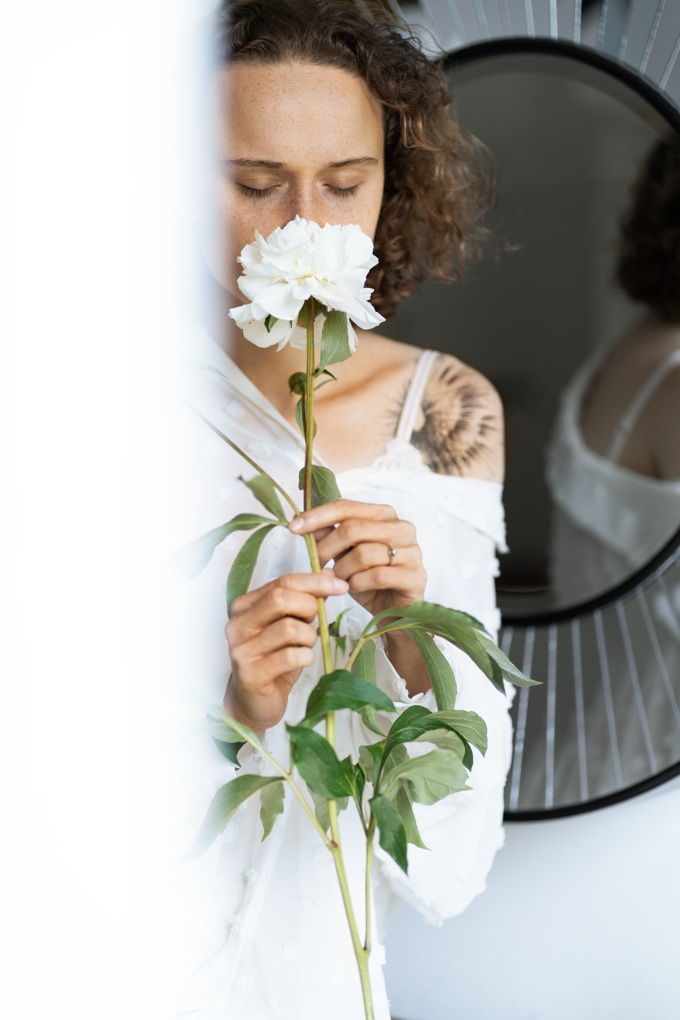 Someone in white holding a delicate white peony flower against a wall with a circular mirror.