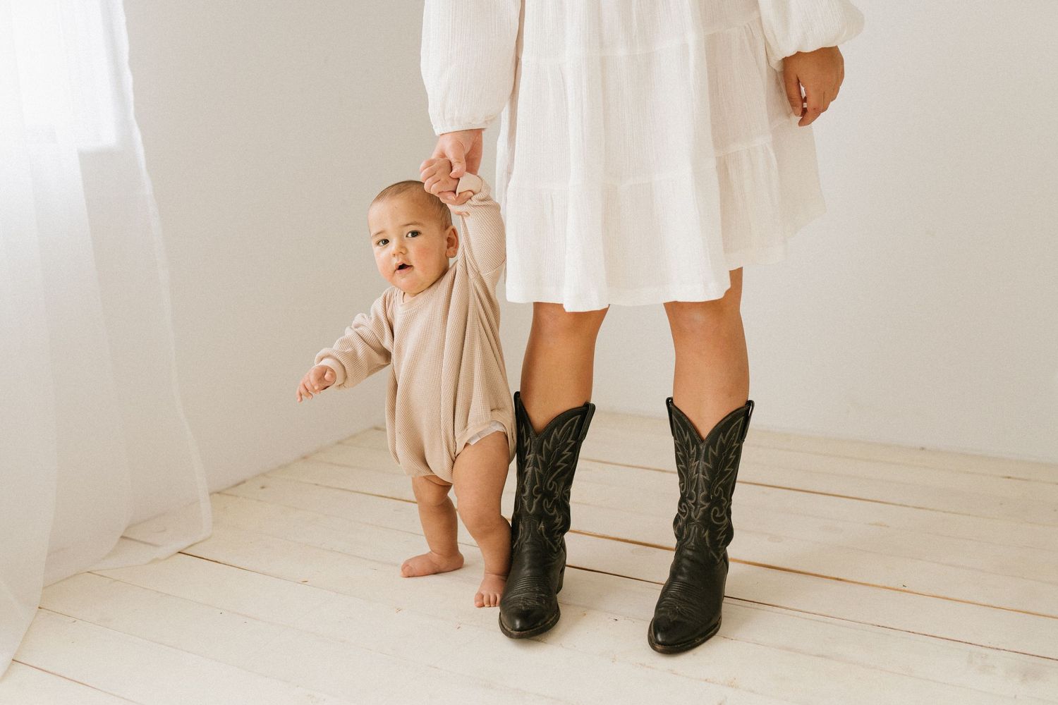Woman in white dress and cowboy boots stands with baby in matching neutral outfit.