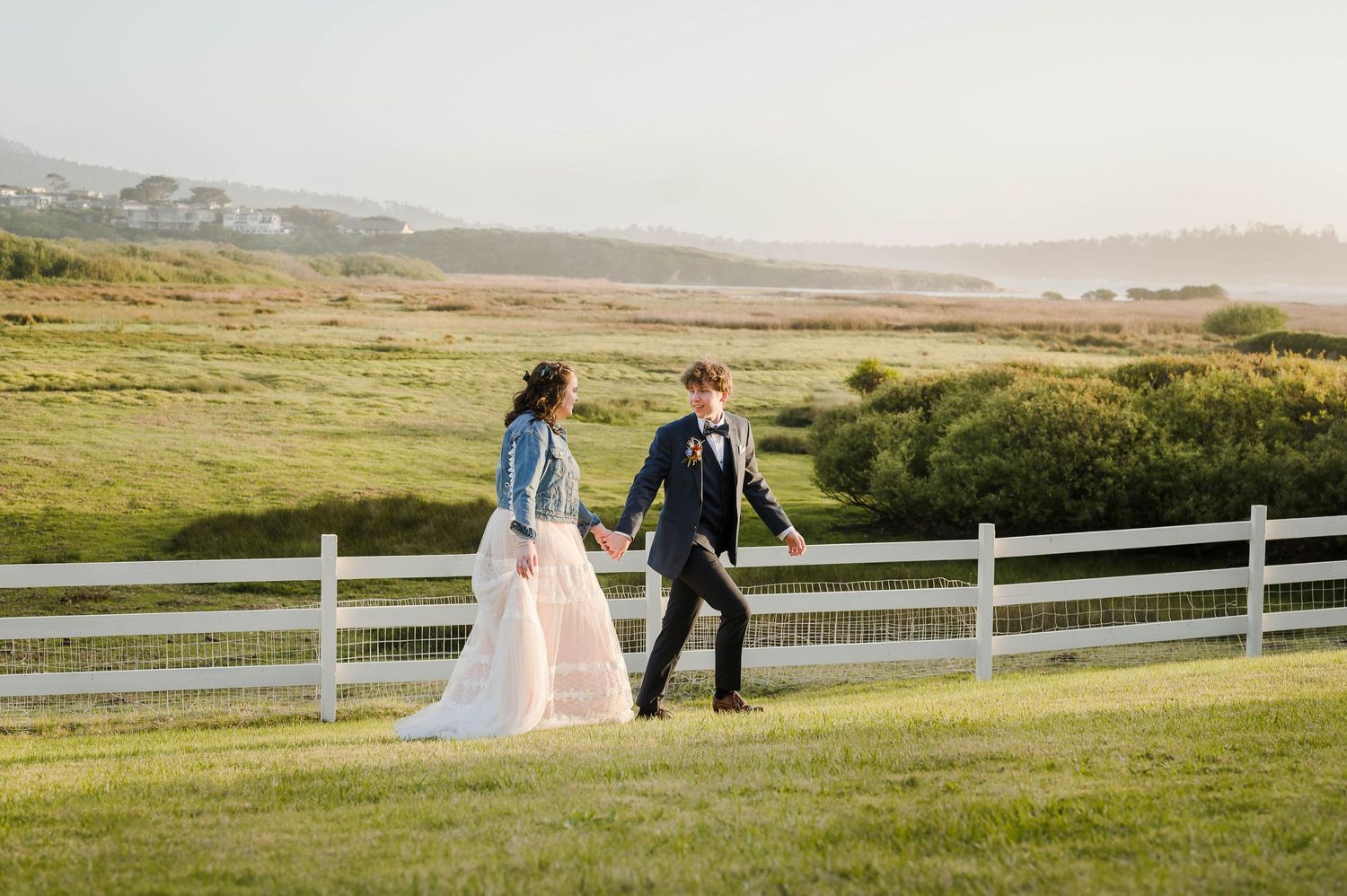 A couple walks hand in hand on grassy hills with white fence and misty landscape.