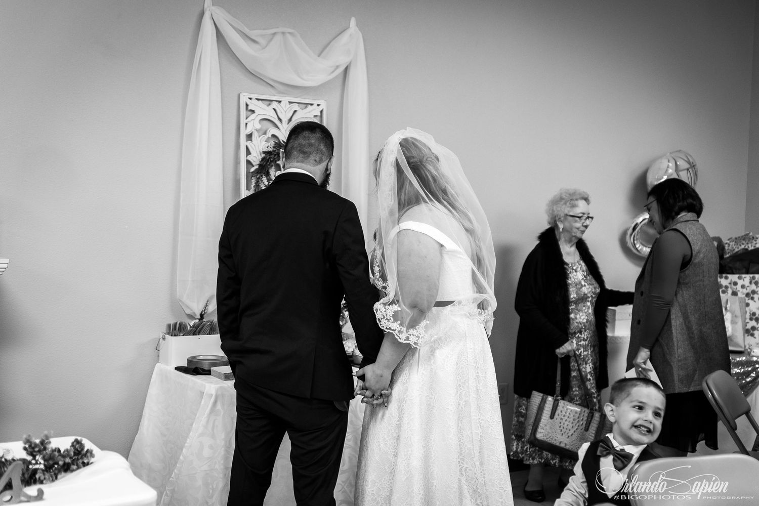 Black and white wedding ceremony photo showing couple and guests gathered around a draped altar.