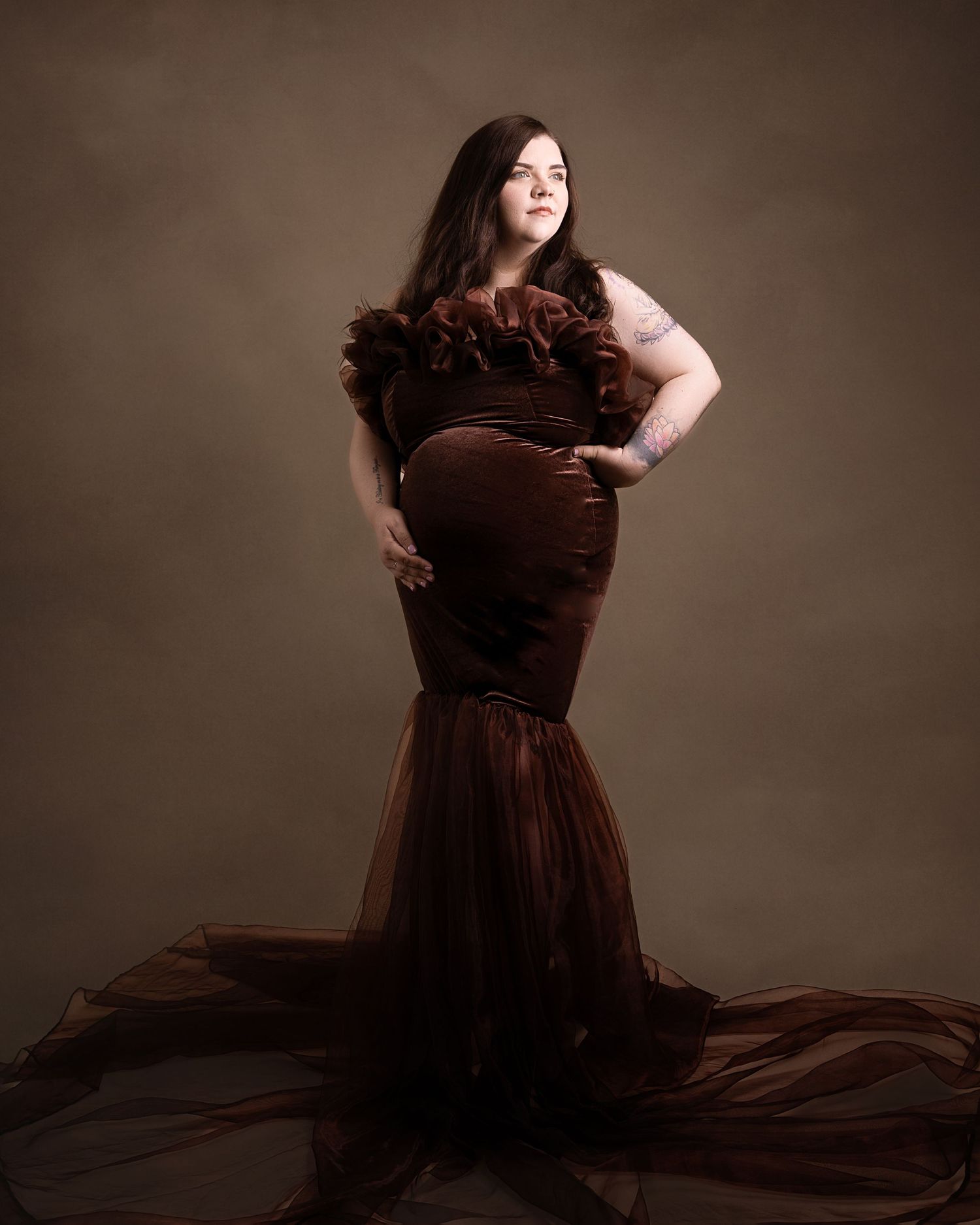 Dramatic maternity portrait in burgundy mermaid gown against brown backdrop.