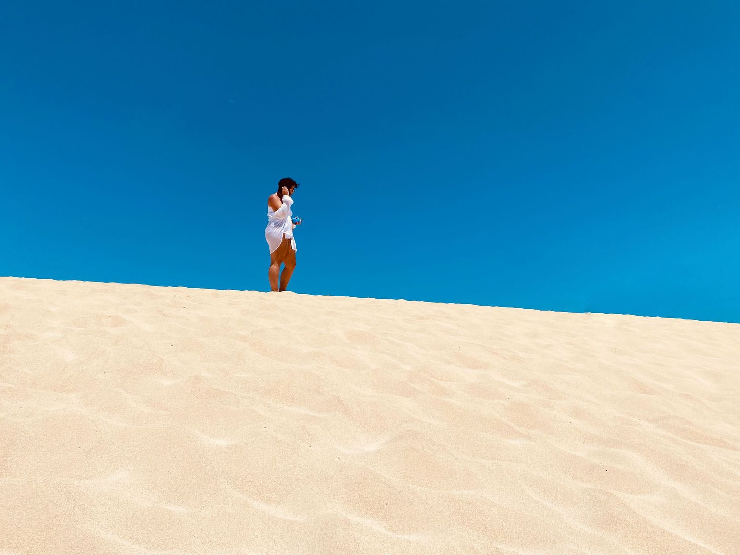 People silhouetted against bright blue sky walking along sandy dune ridge.