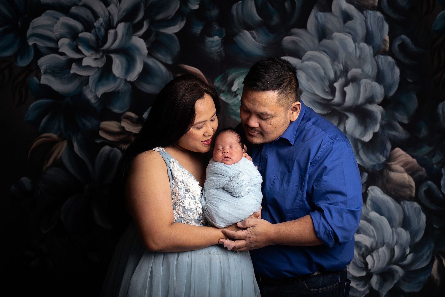 Family portrait against dark floral backdrop wearing coordinated blue and white outfits.