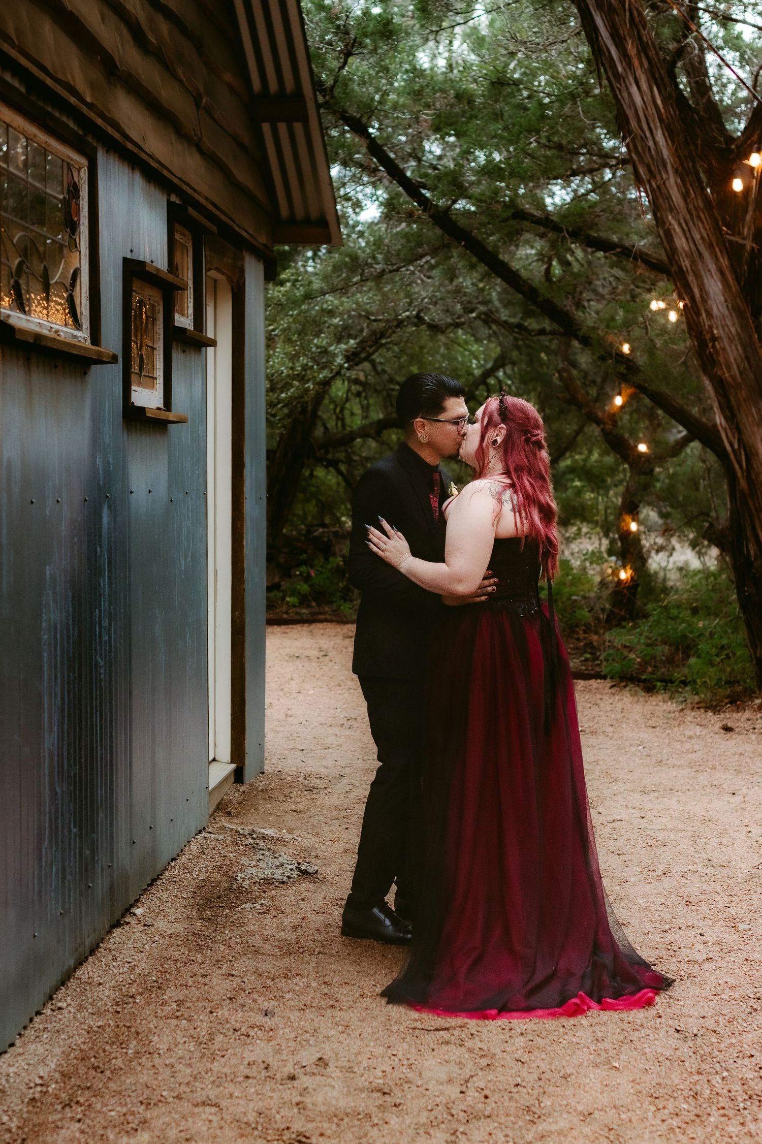 Romantic couple embracing outside rustic building with string lights and forest backdrop.