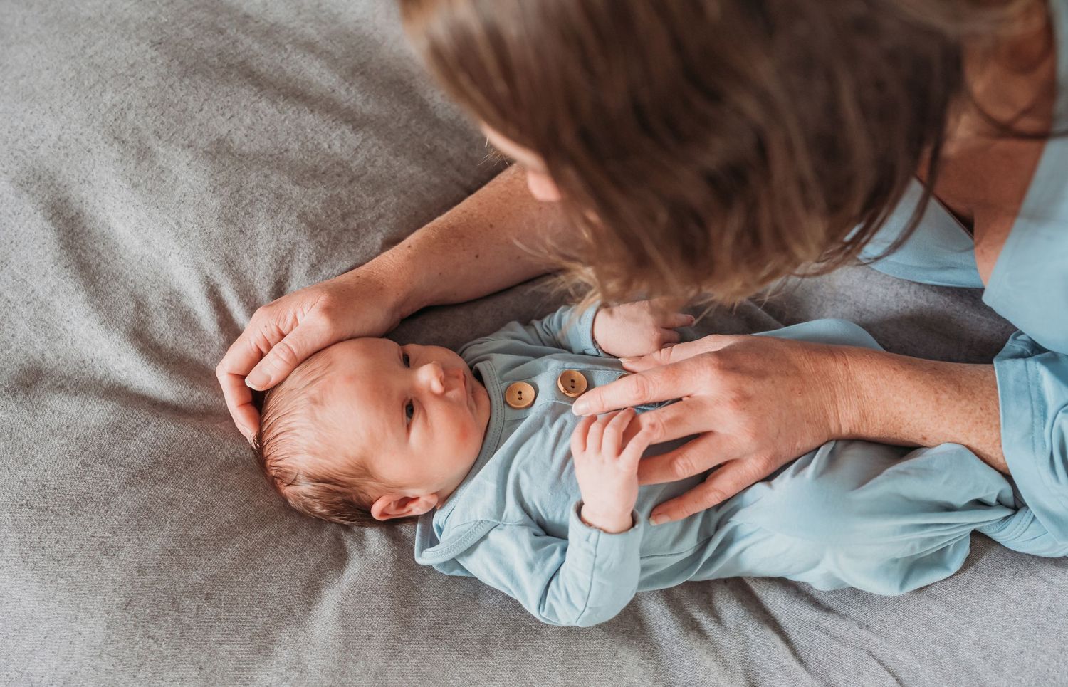 Newborn baby boy lays on gray comforter looking up at mom.