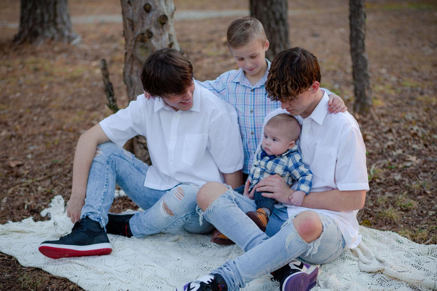 Three brothers in white shirts and jeans sit on a blanket outdoors, lovingly holding their newborn baby sibling.