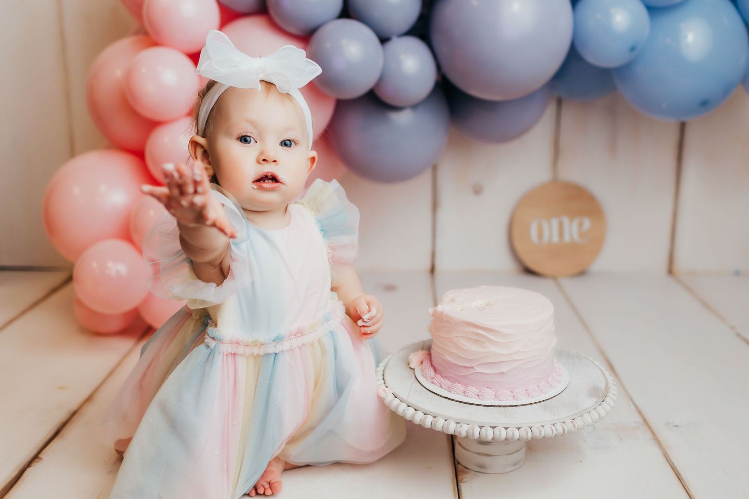 Baby in pastel dress celebrates first birthday with pink and blue balloon arch and cake.