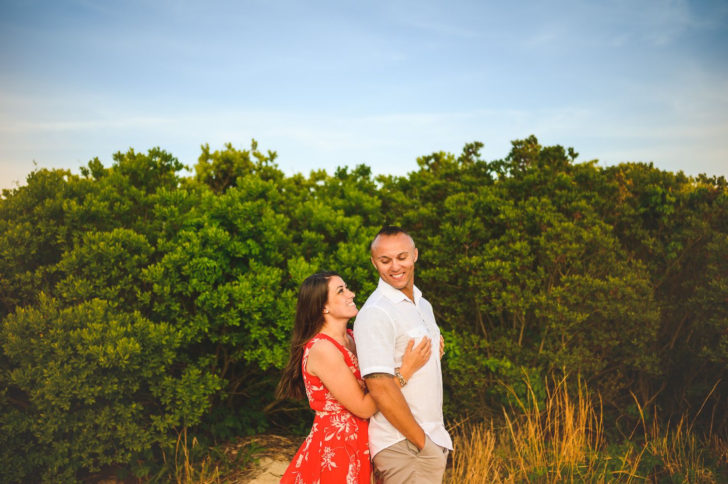 engaged couple on LBI beach smiling and holding each other in front of green bush
