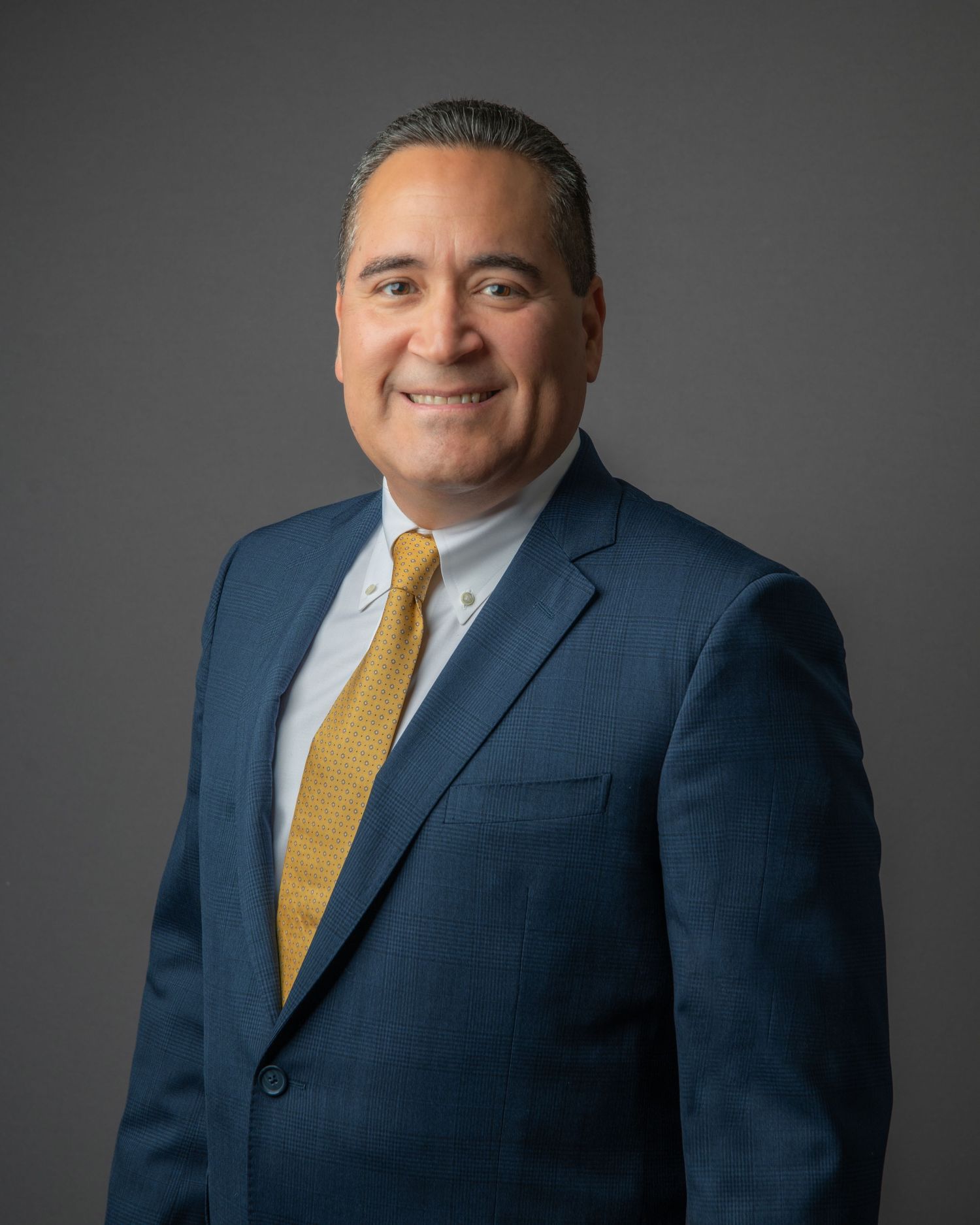 Professional headshot of man in navy suit with yellow tie against grey background.