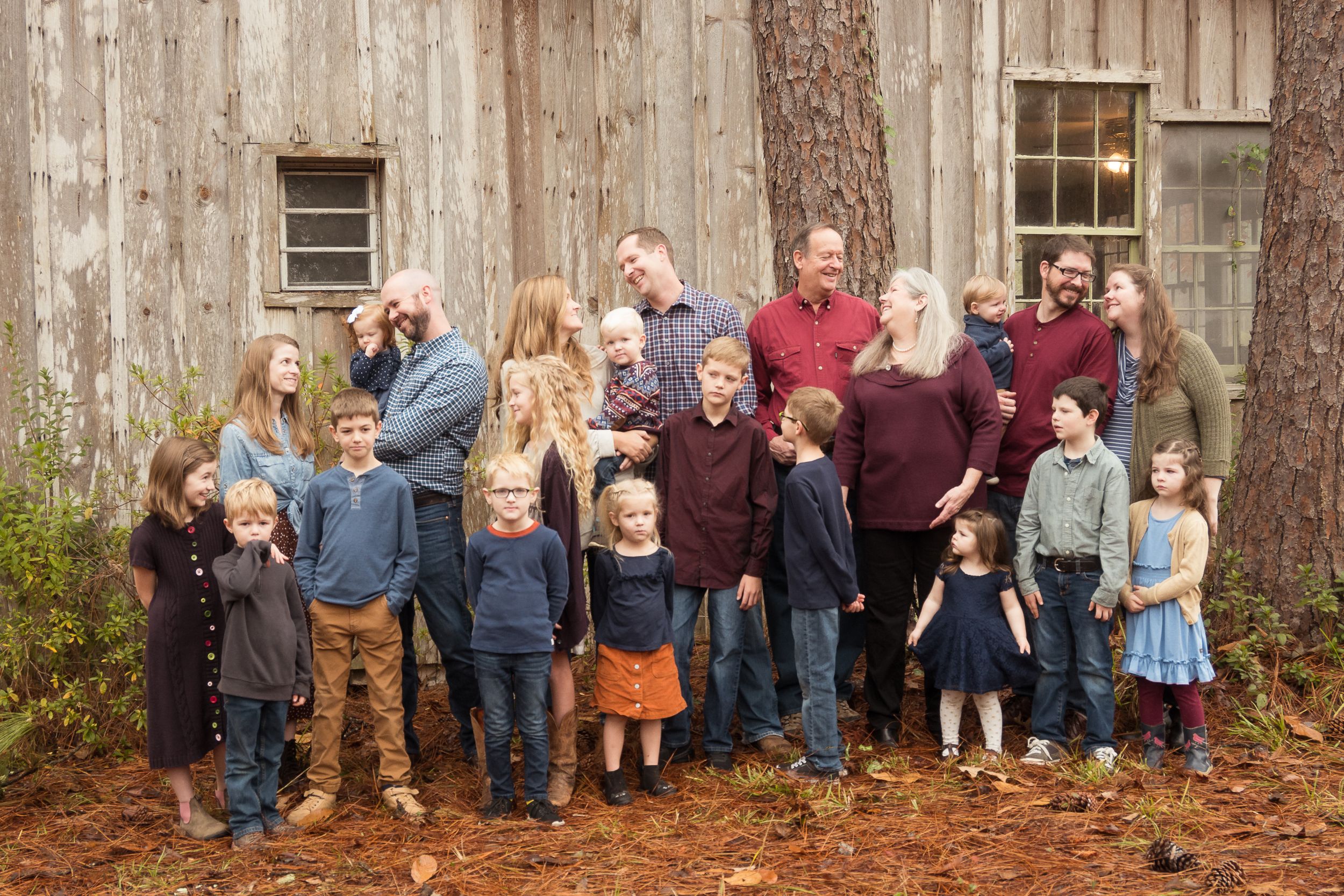 A large family group poses together outside a rustic wooden barn on fallen autumn leaves.