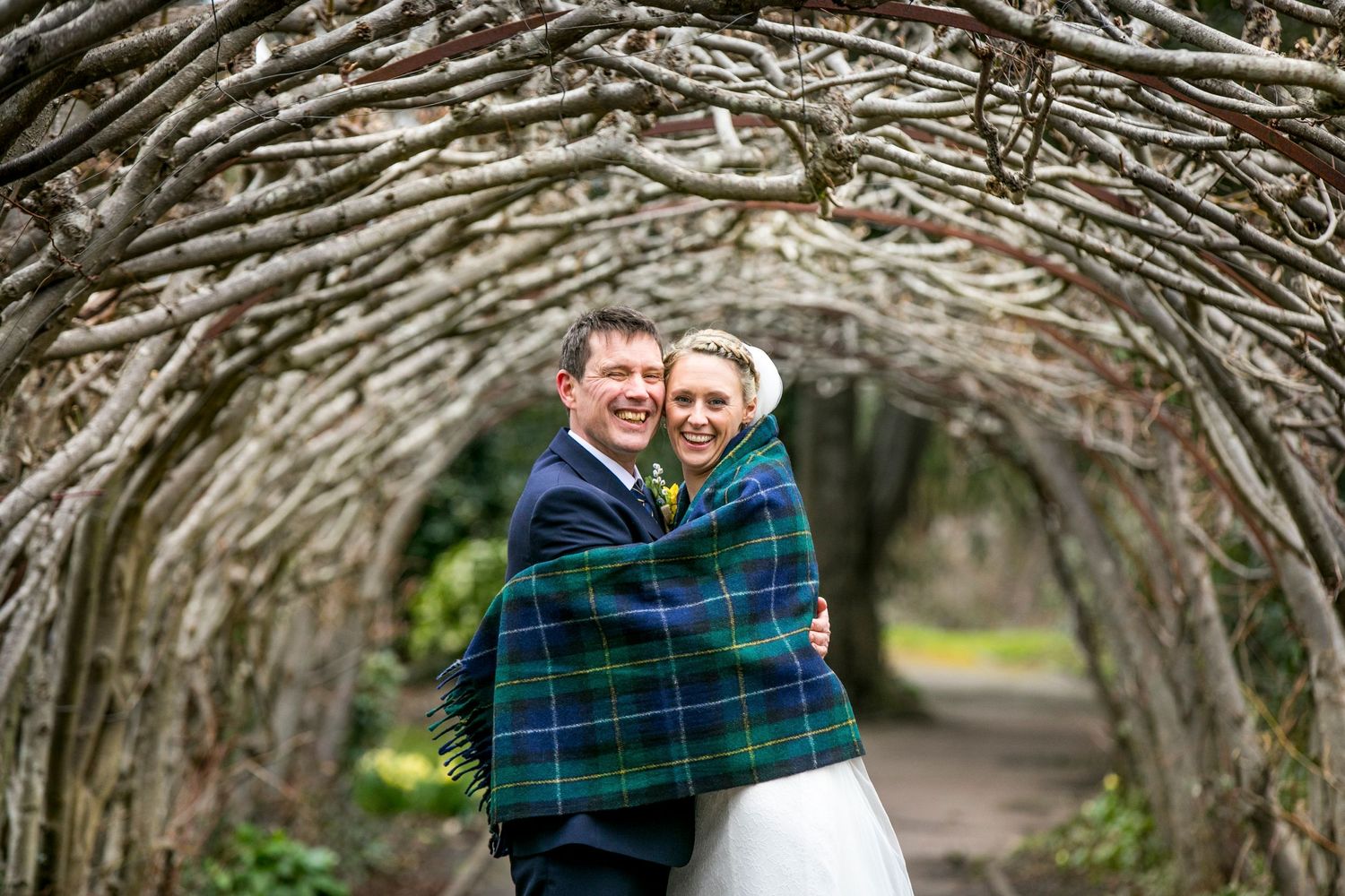 Couple embracing under a natural archway of intertwined branches in a garden setting.