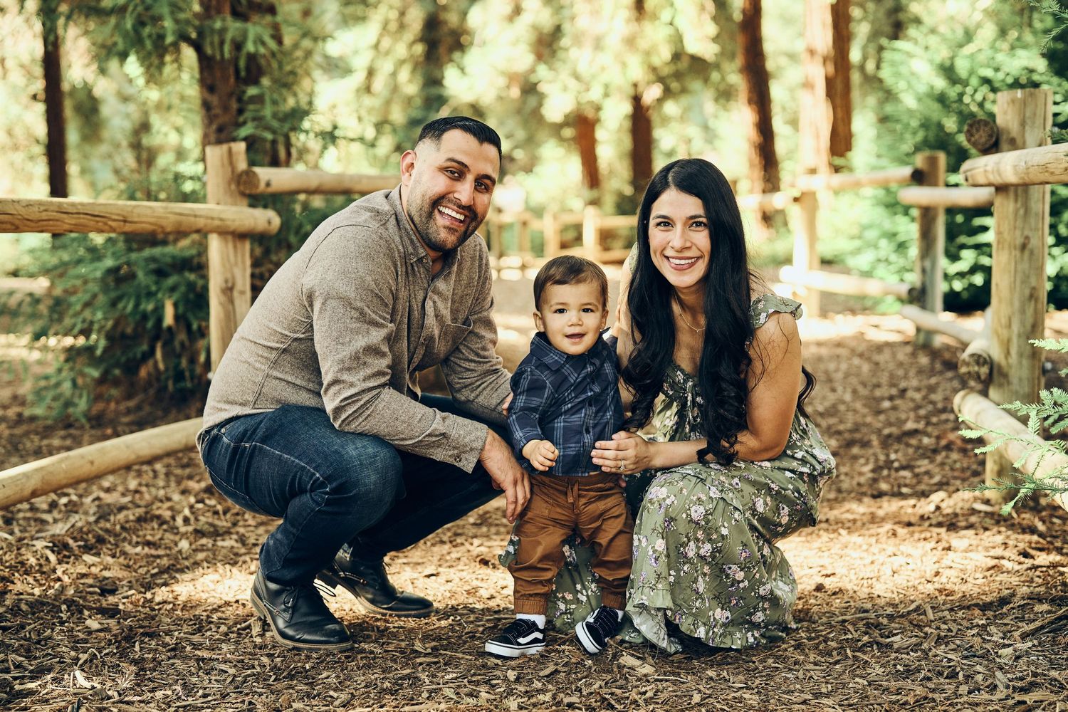 Family of three posing together on wooden pathway in forest setting.