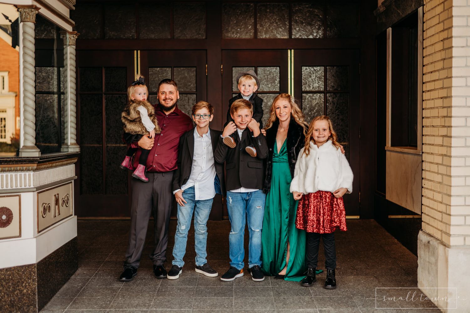 Large family of seven poses together smiling outside ornate building doors during holiday photo session.