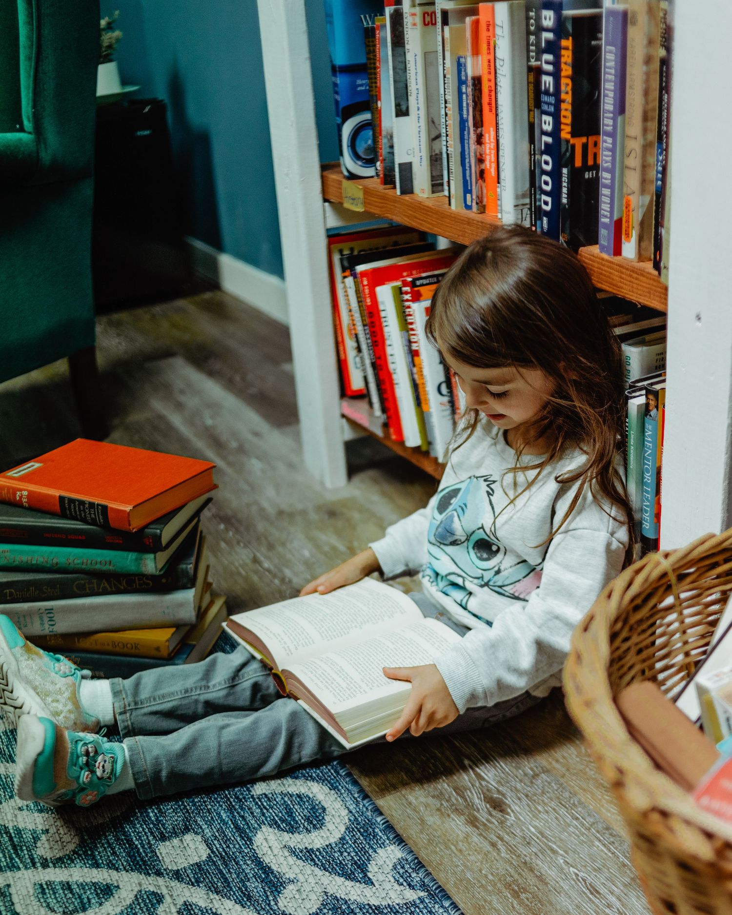 Child in light blue sweater reading book while sitting on patterned floor near bookshelf.