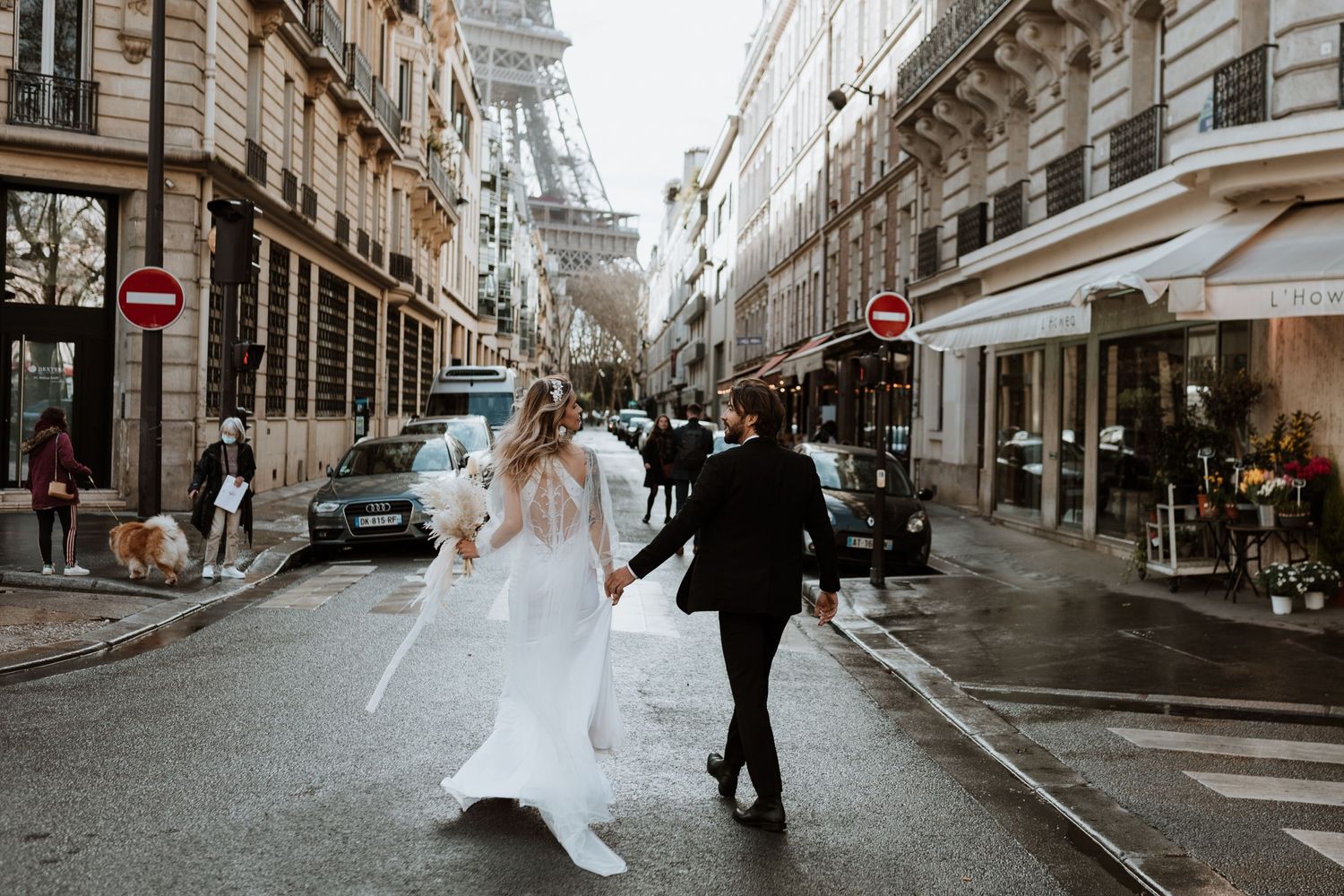 A couple in wedding attire walking down a charming Paris street with the Eiffel Tower visible in the background.