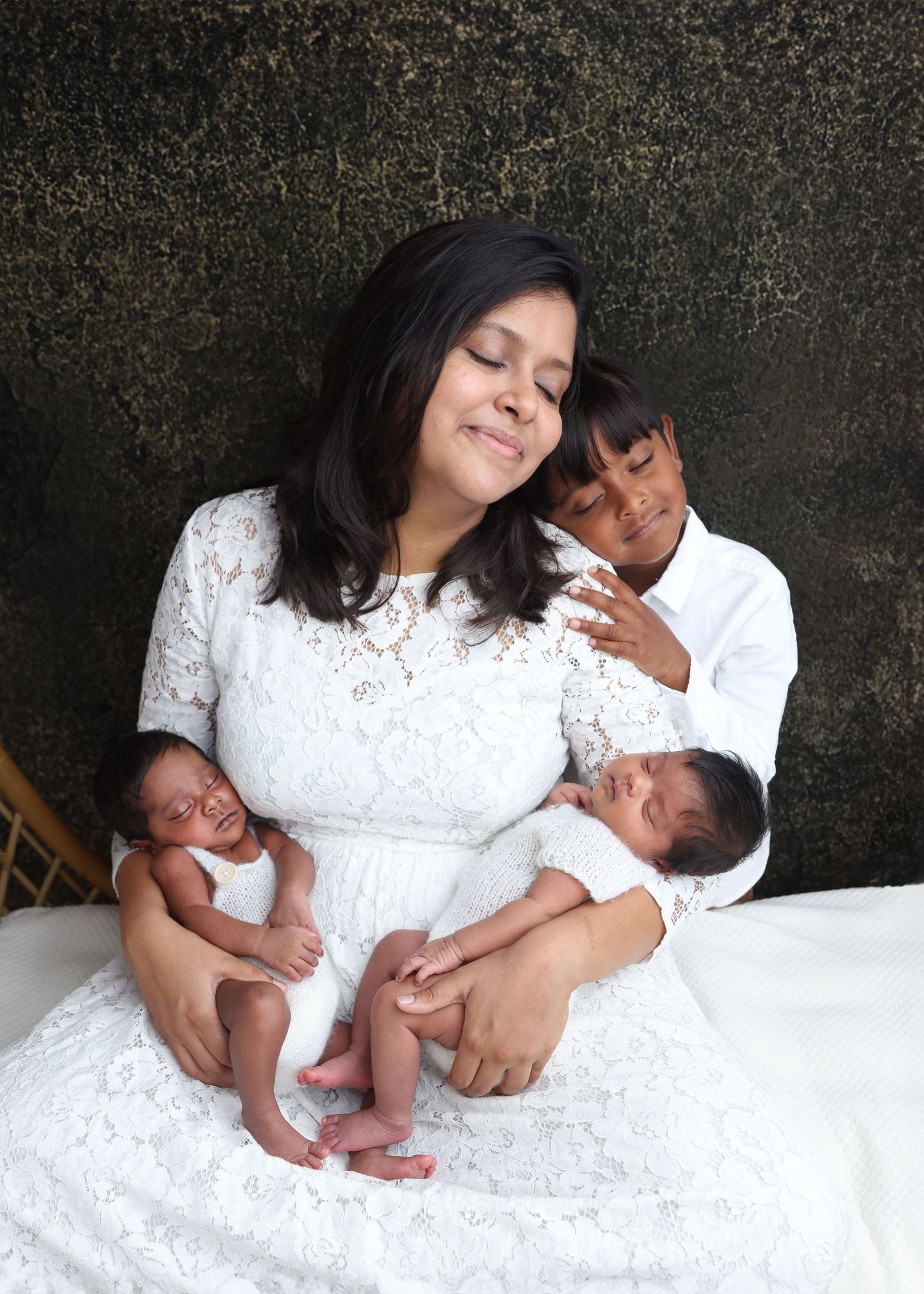 Tender family portrait with newborn twins in white lace setting against textured backdrop.