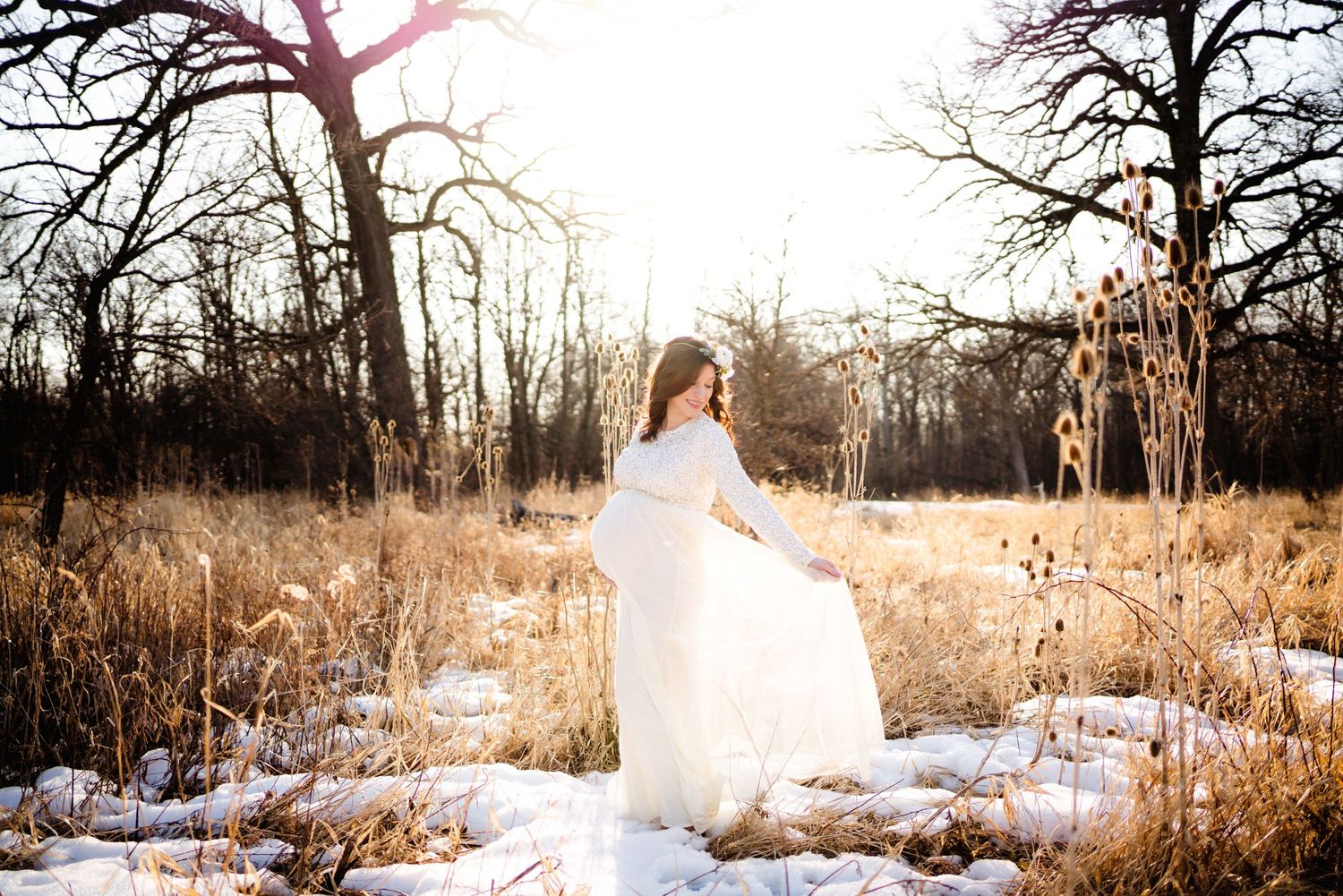 Woman in white dress twirling in snowy field with bare trees during winter photoshoot.