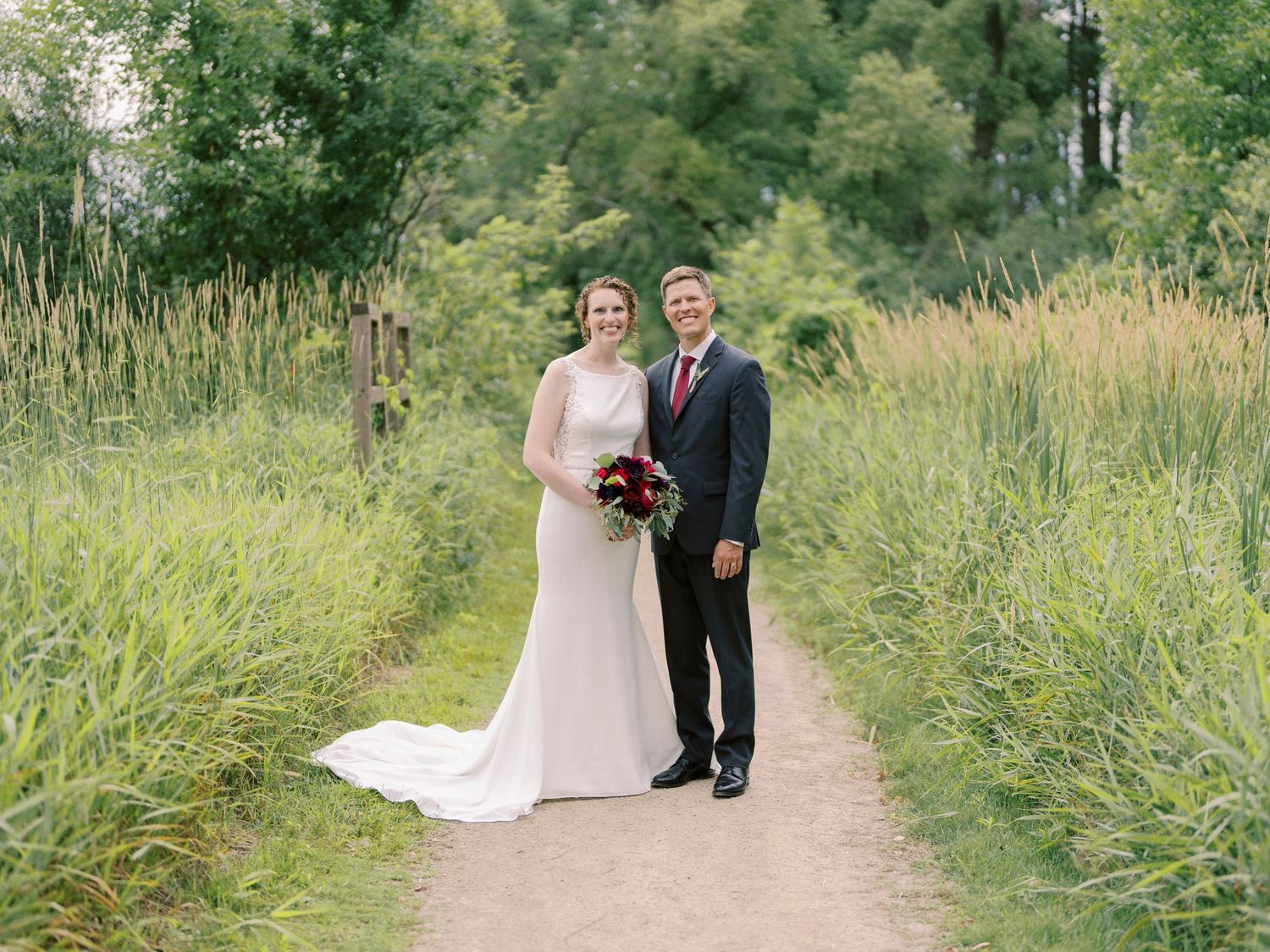 A bride and groom pose together on a nature path surrounded by tall greenery and wildflowers.