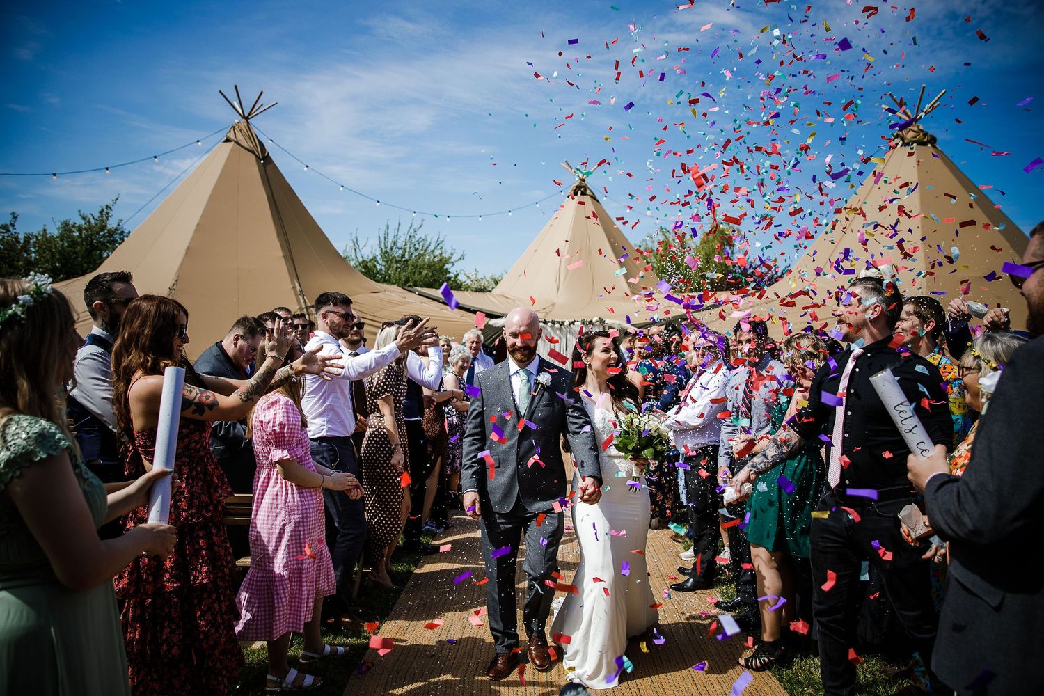 Celebration with pink and purple confetti being thrown in the air near outdoor tipi tents on a sunny day.