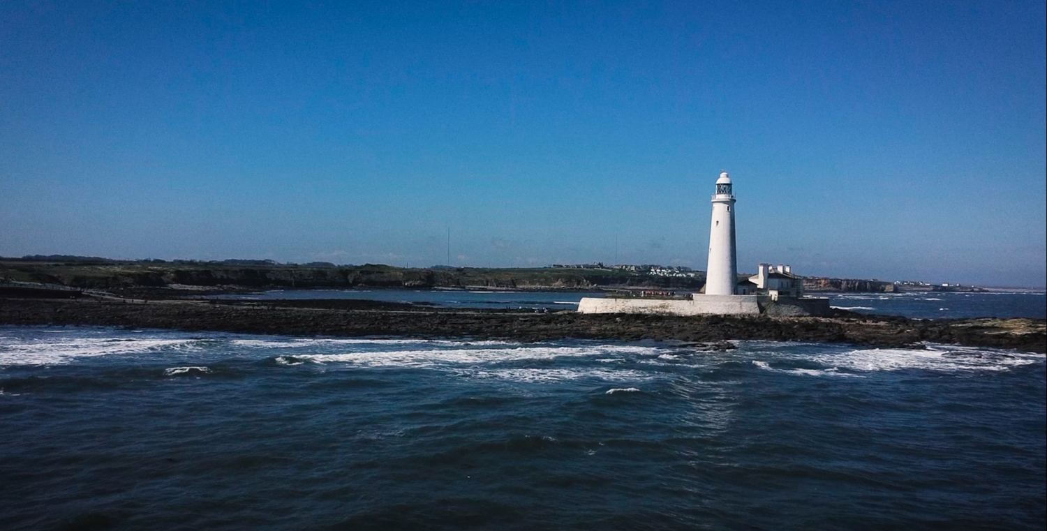 A white lighthouse stands on rocky shores against blue ocean waves.