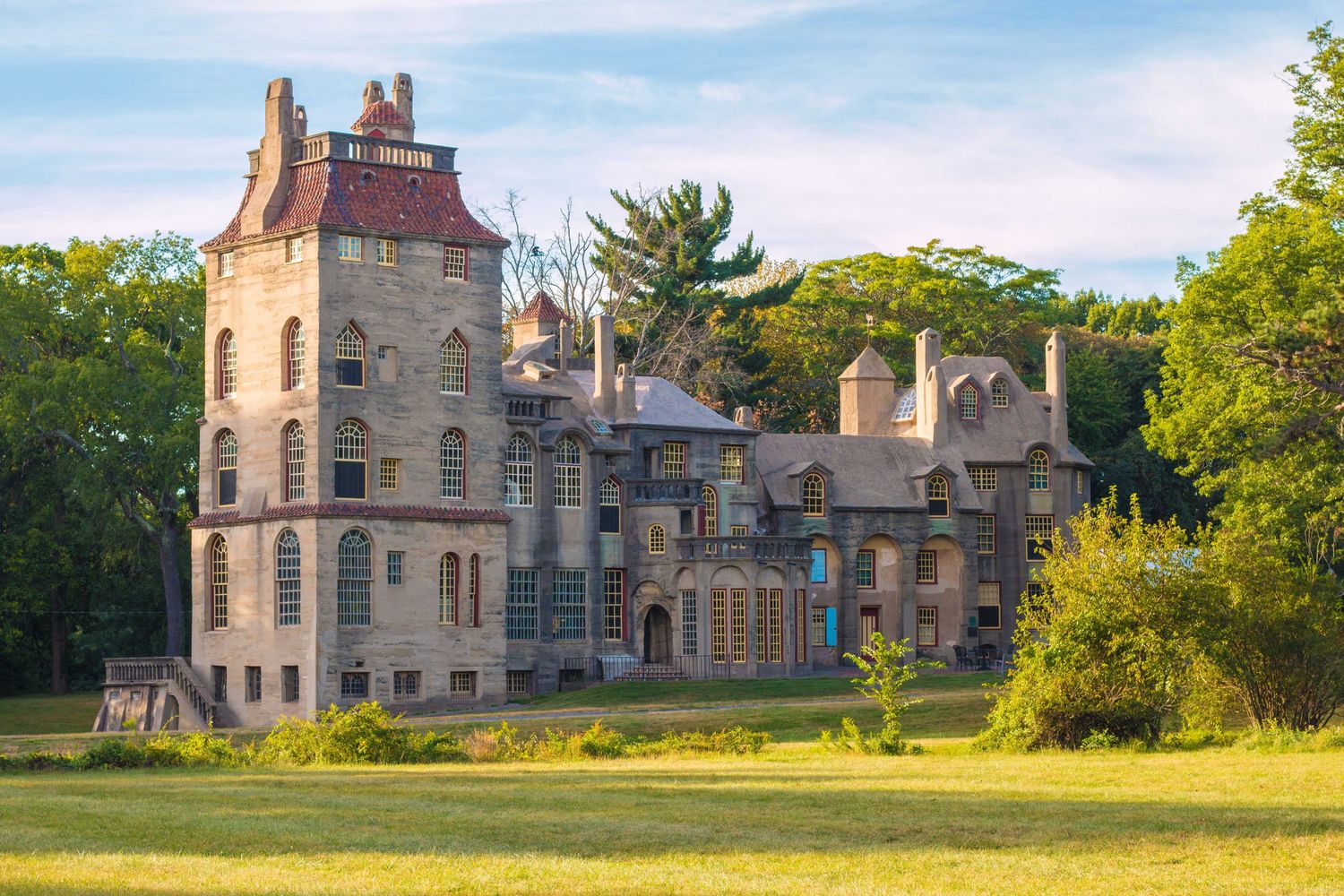 A grand castle-like mansion with stone towers and turrets stands surrounded by green trees at sunset.
