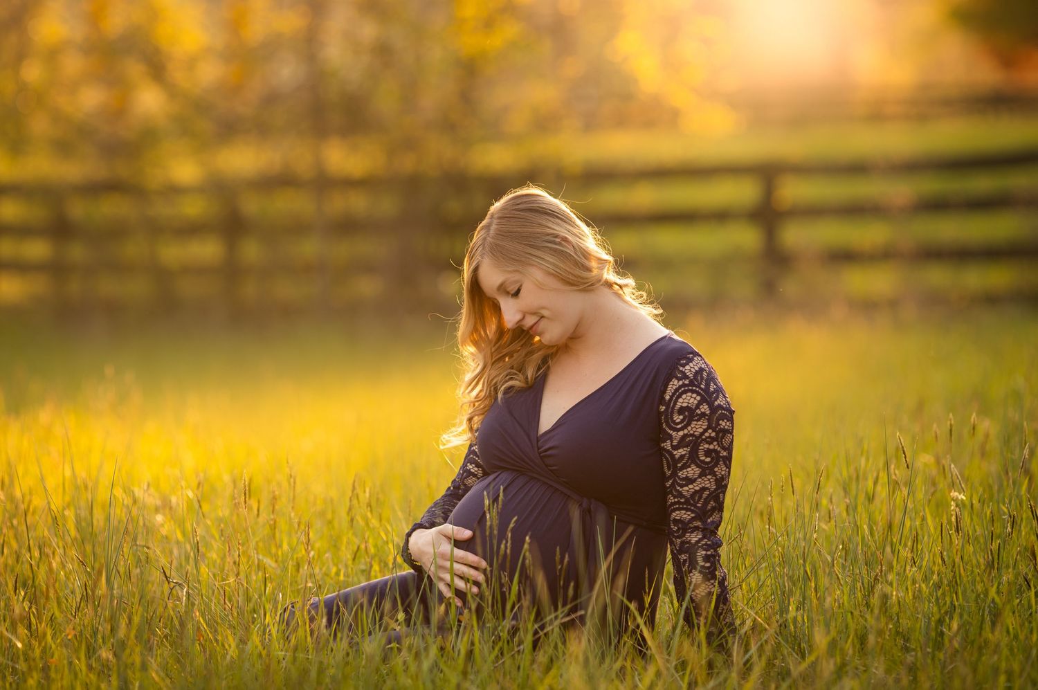 Maternity photo of expectant mother in navy dress sitting in sunlit field.