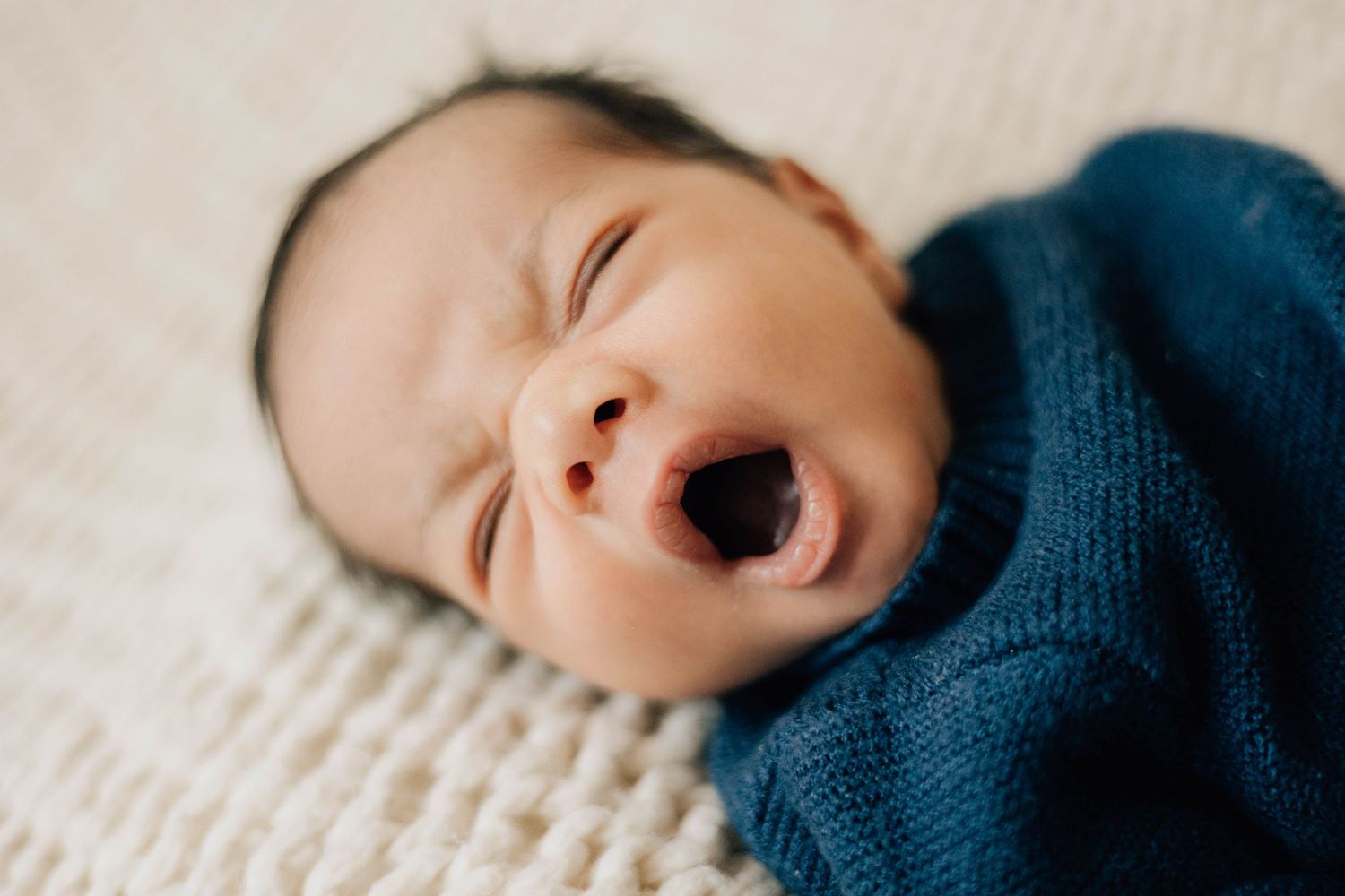 Newborn baby wrapped in a blue knit swaddle yawning while lying on a cream textured blanket.