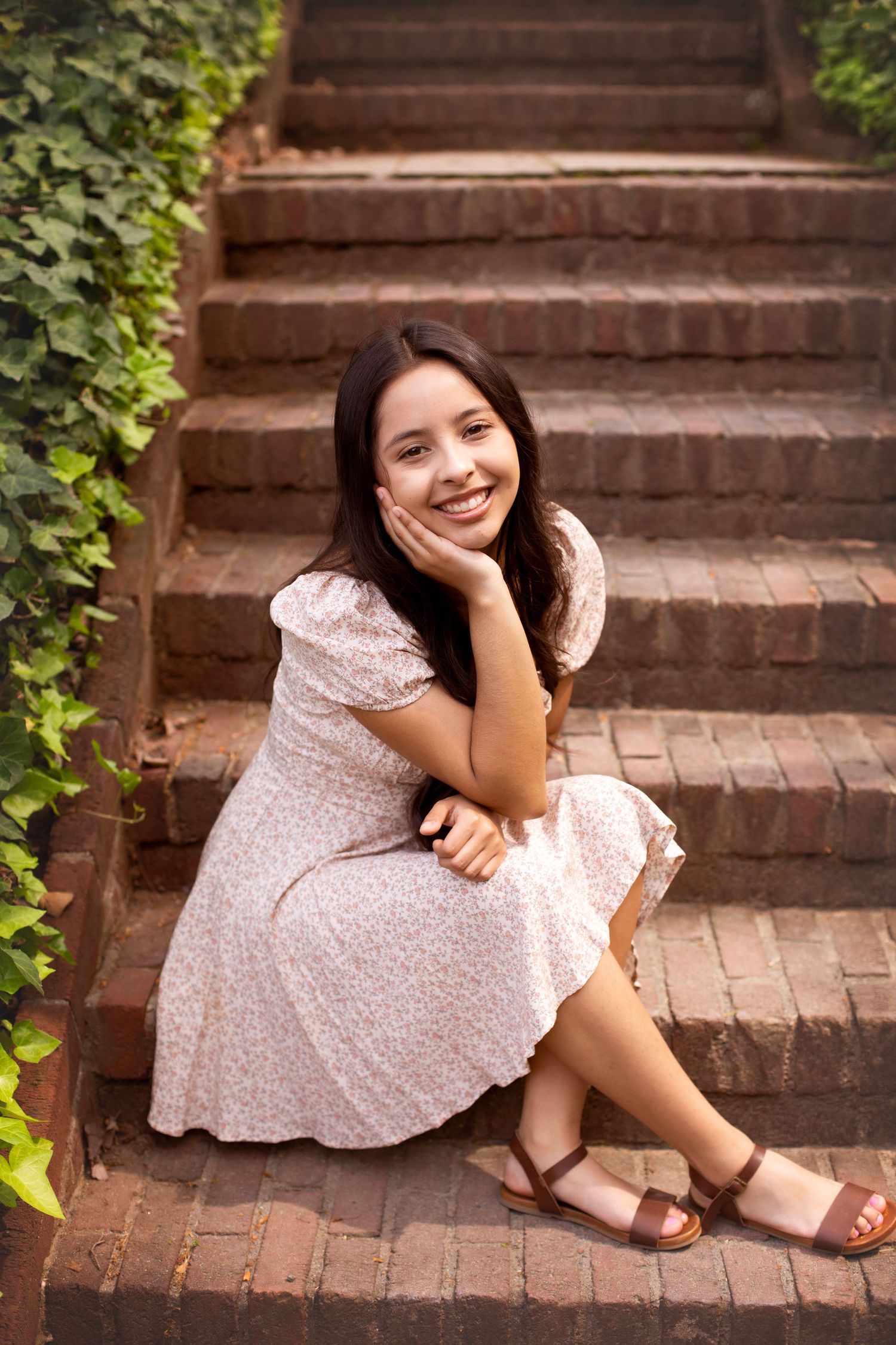 High school senior wearing a floral dress, sitting on bricks, ivy growing up the bricks behind her.