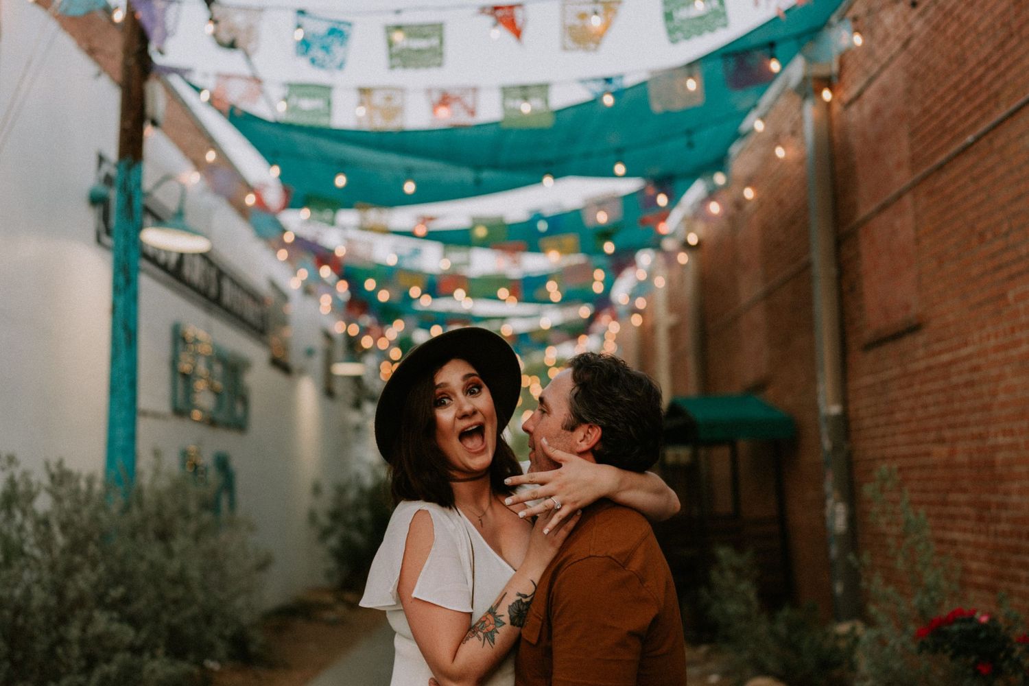 A romantic couple shares a joyful moment under festive string lights and colorful banners in an alley.