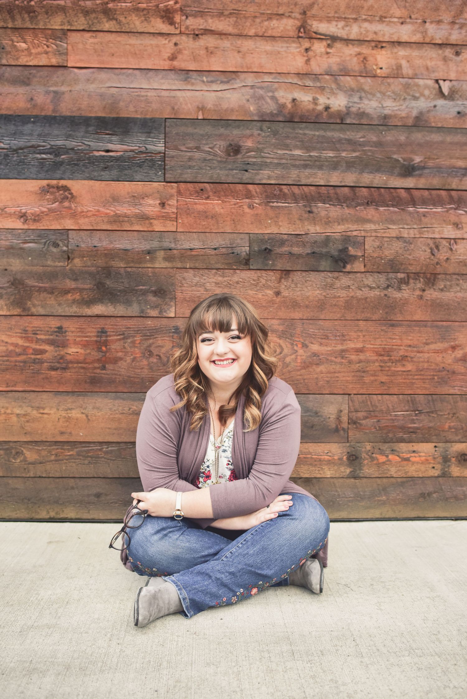 Person in casual attire sits cross-legged against rustic wooden wall backdrop.