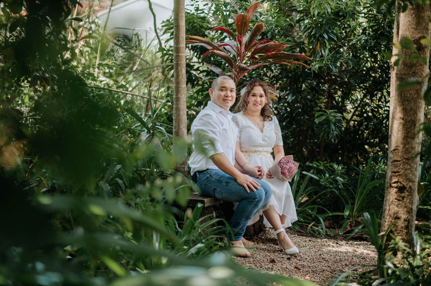 A couple sits together on a garden bench surrounded by lush tropical greenery in an outdoor setting.