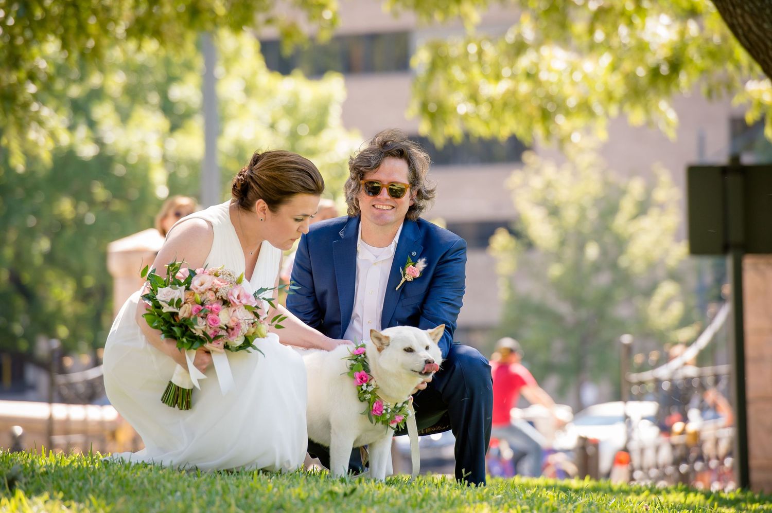 A couple and their white dog share a moment on a sunny lawn with pink flowers and trees in the background.