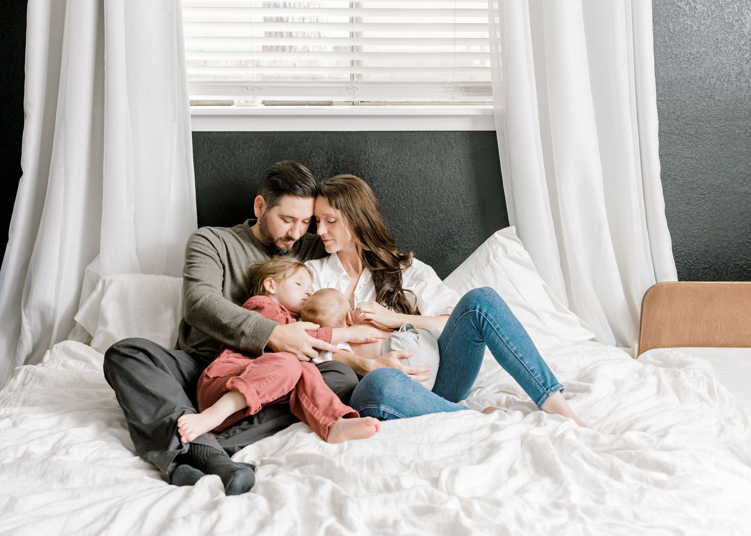 family of four cuddled together in their bed. husband and wife's heads are together as big sister kisses little brother