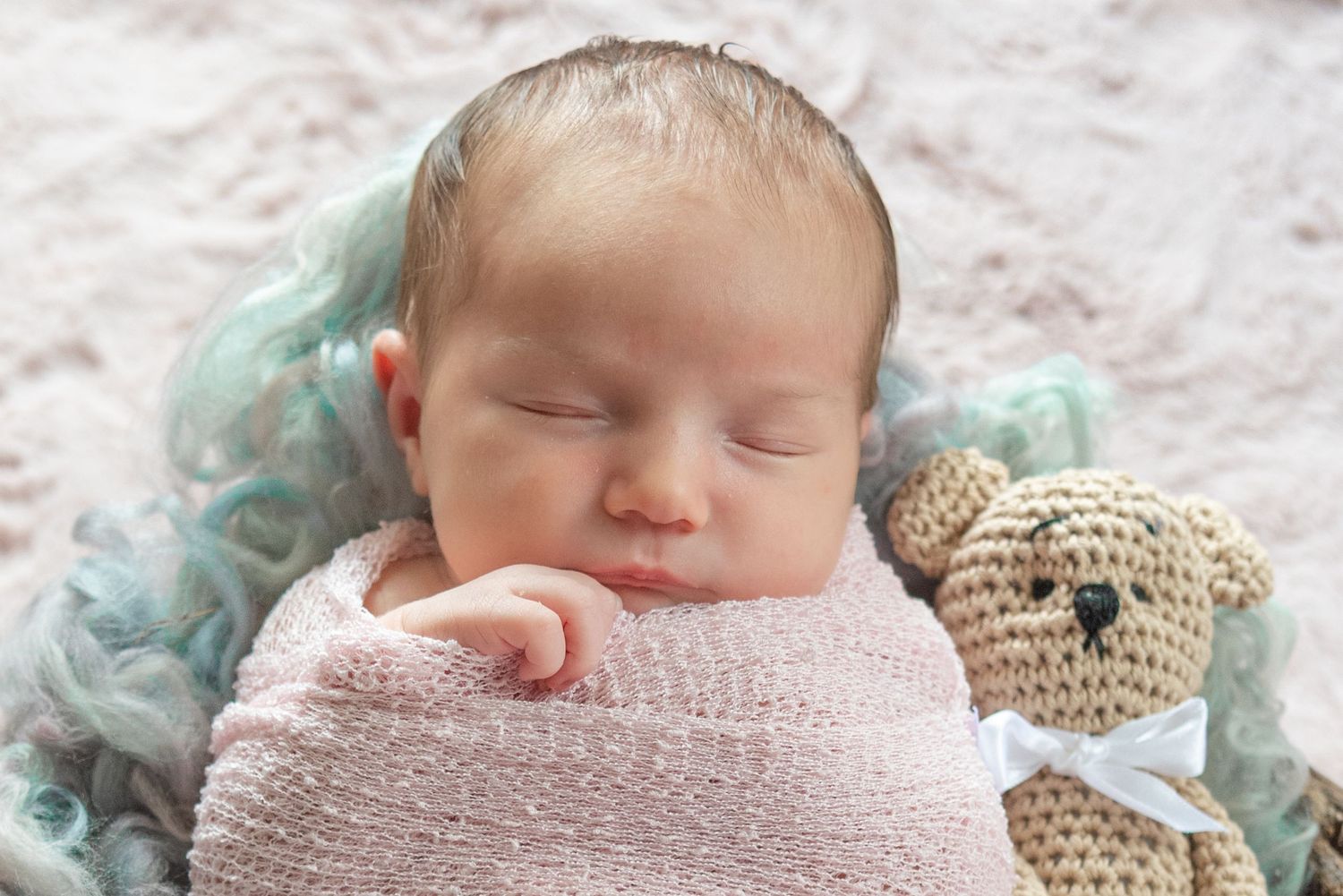 Peaceful sleeping newborn swaddled in pink with mint accents and stuffed bear companion.
