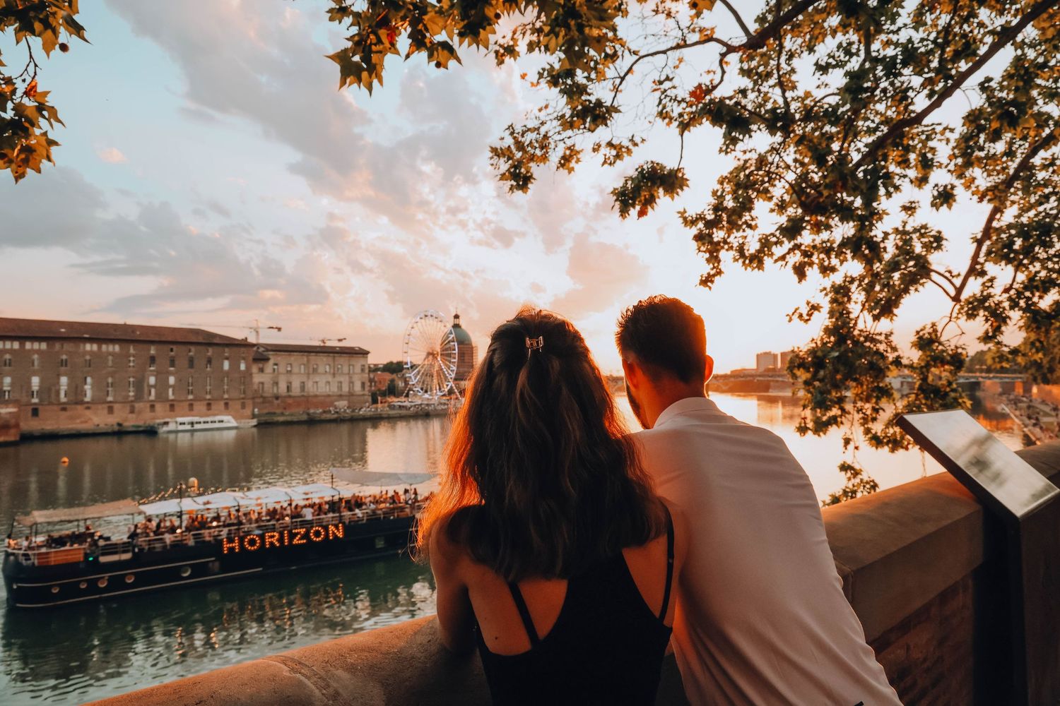 Silhouetted figures watching sunset over a scenic river with boats and historic architecture.