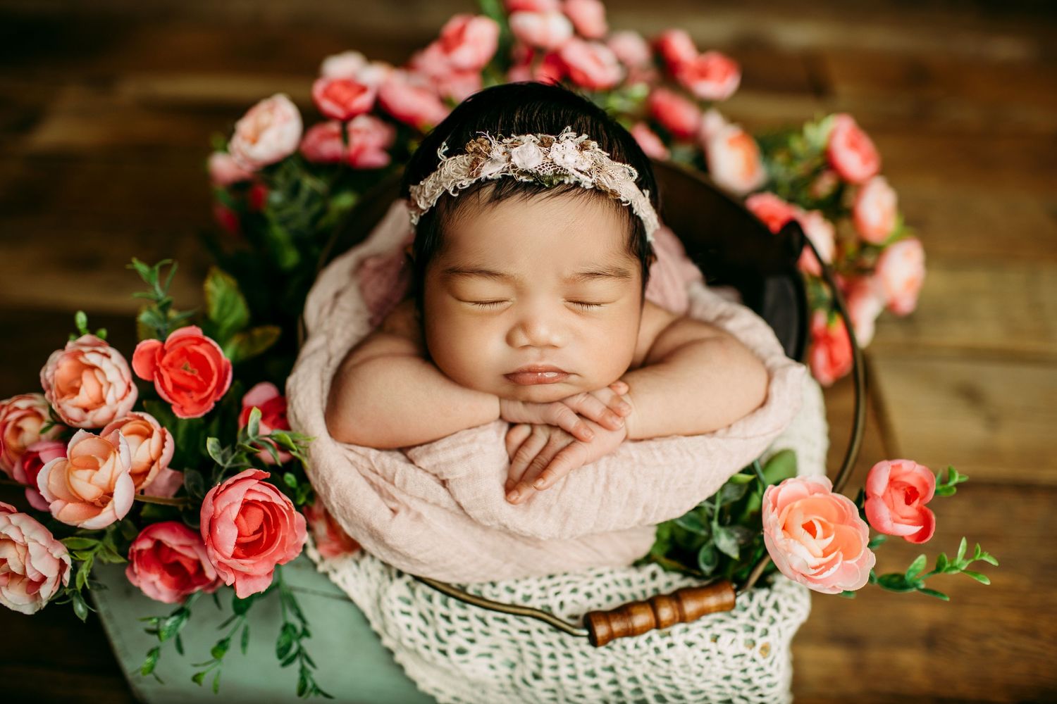 Newborn baby sleeping in basket surrounded by pink roses and greenery.