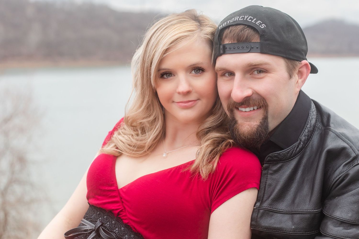 A couple poses together near a lake on an overcast winter day.