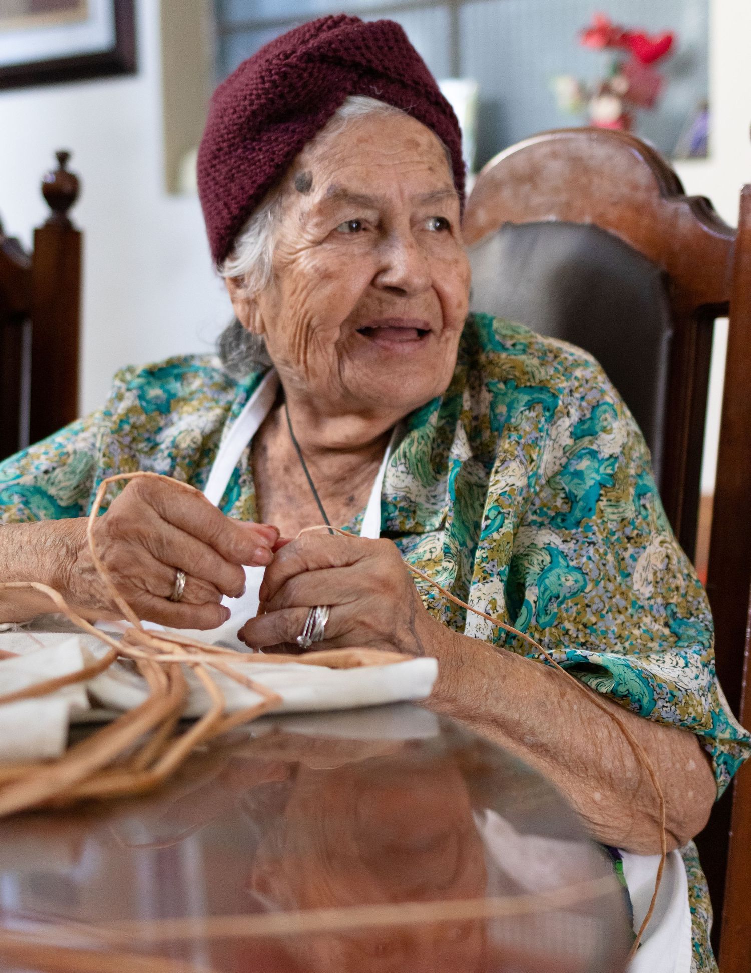 Person in burgundy hat and floral dress writing at wooden table indoors.