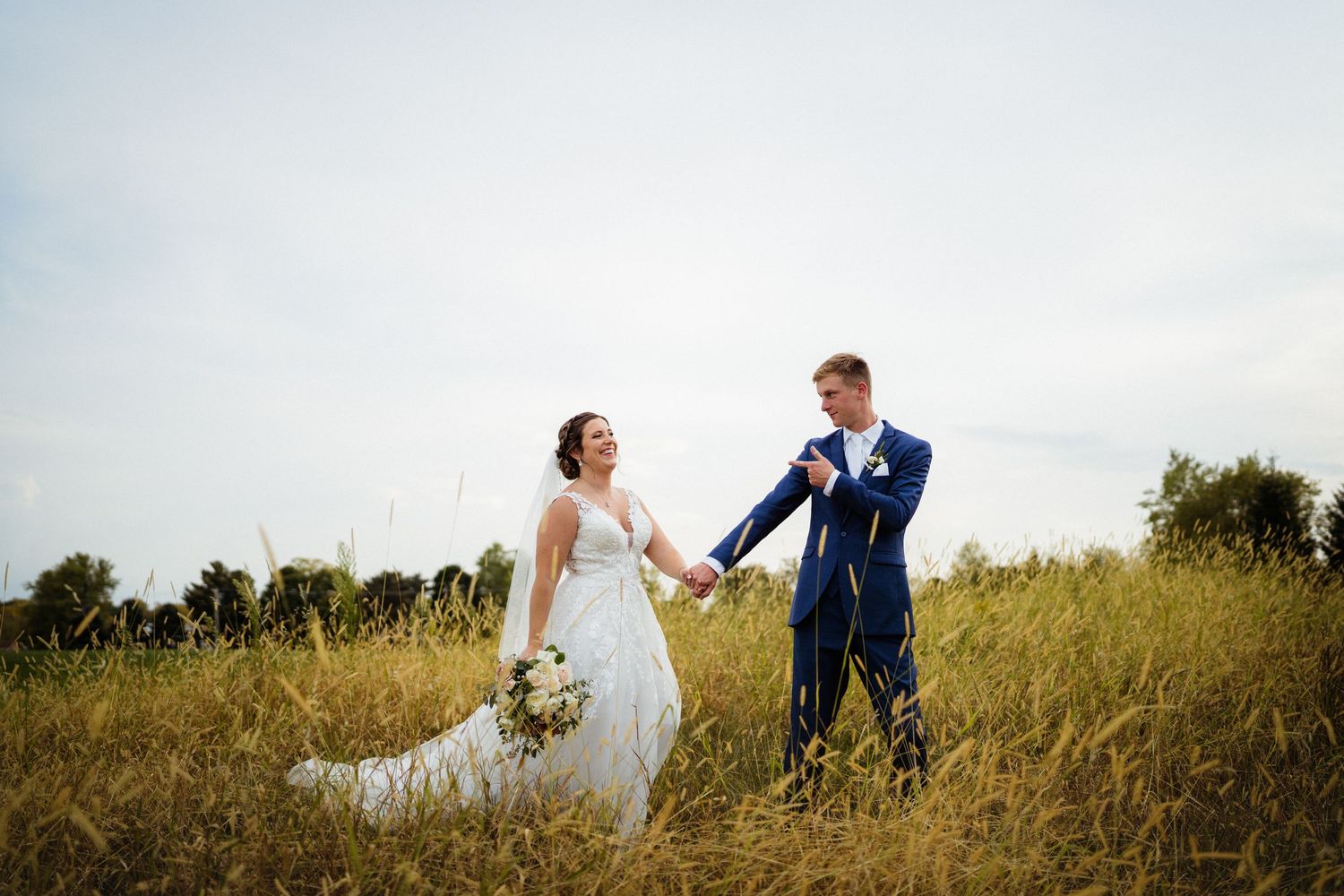 Wedding couple holding hands and walking through a golden field under cloudy skies.