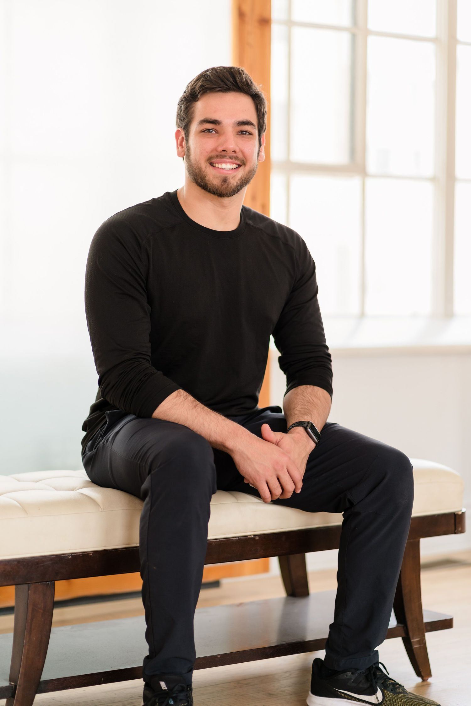 Professional portrait of person in black sweater seated on light colored bench in bright room.