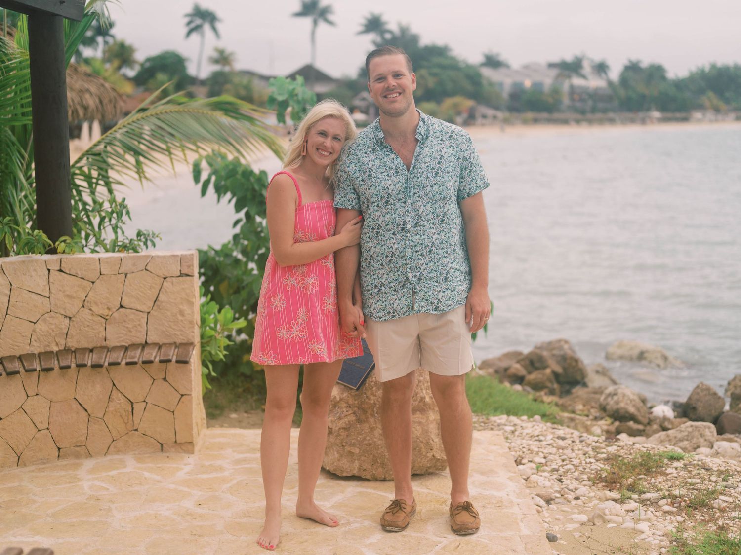 A couple stands on a stone patio overlooking a tropical waterfront with palm trees and rocks along the shoreline.