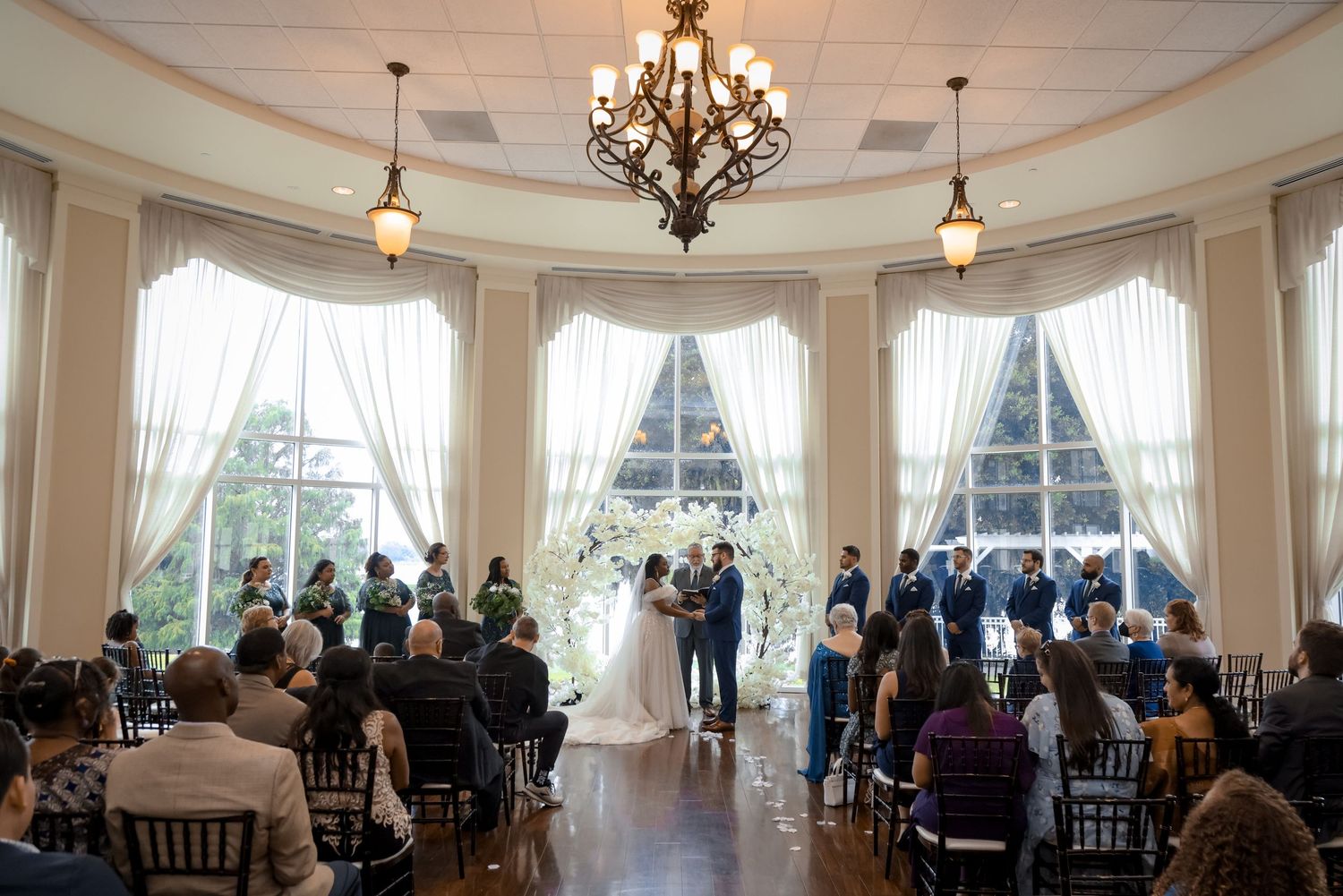 Wedding ceremony in an elegant room with large windows and chandelier.