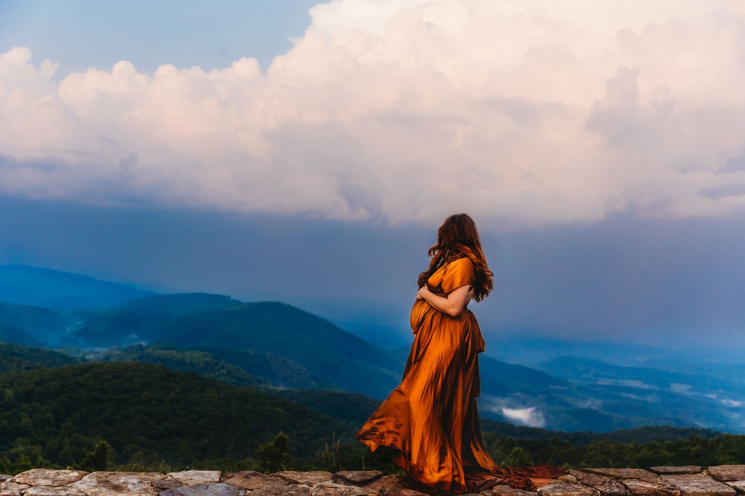 Woman in flowing orange dress stands on mountain peak against dramatic clouds.