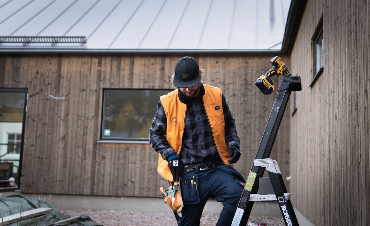 Hantverkare i orange väst förbereder arbete vid stege utanför träbeklädd byggnad. Fotograf Gabriel Alenius, Falun