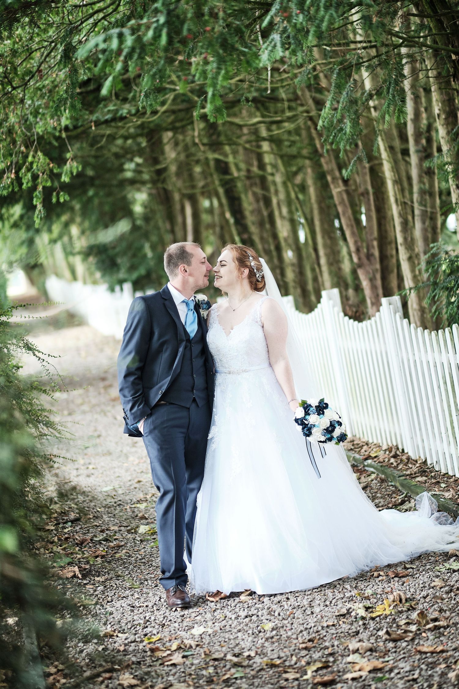 Newlyweds share intimate moment on tree-lined path with white picket fence.