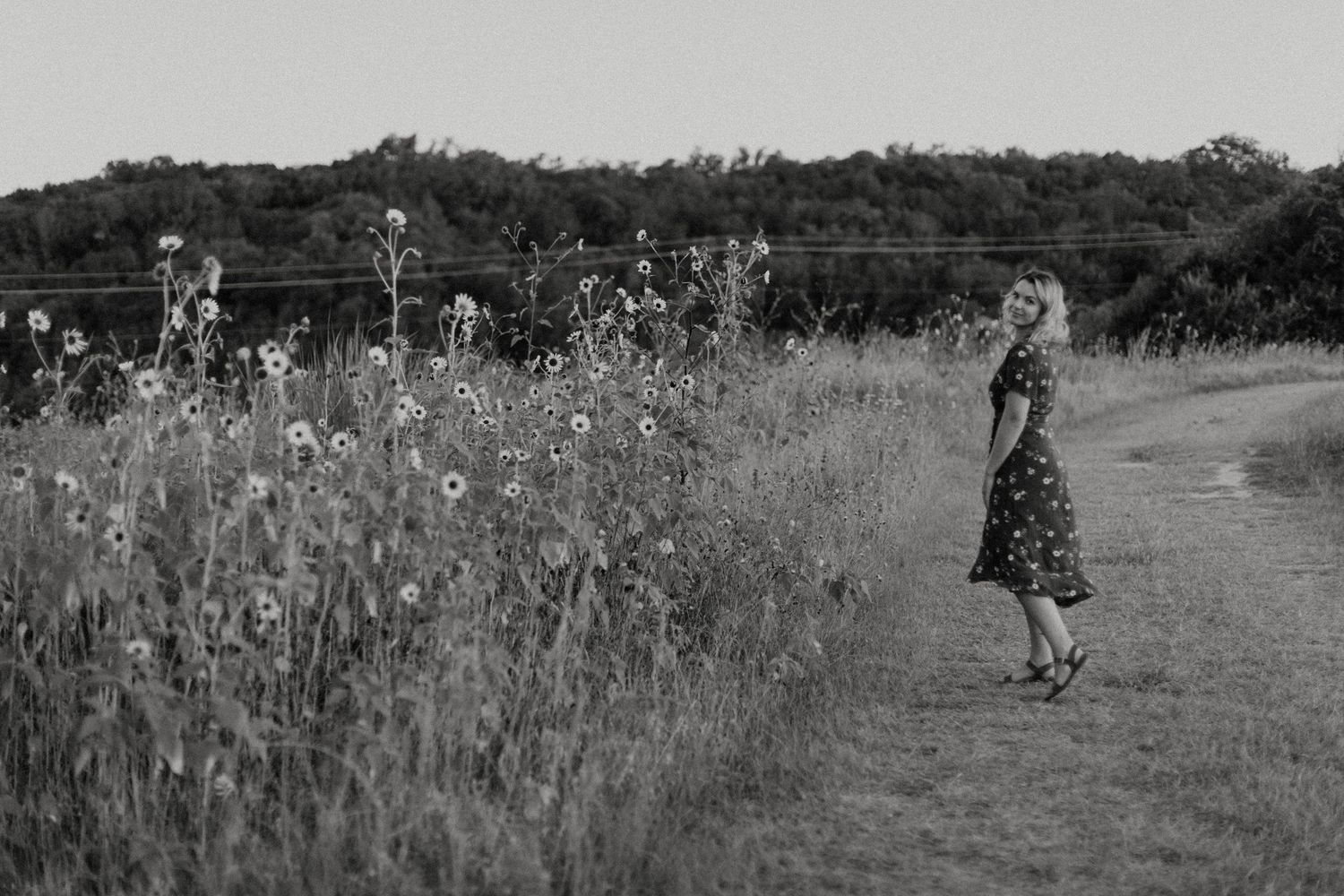 Black and white photo of woman standing among tall wildflowers on rural path.