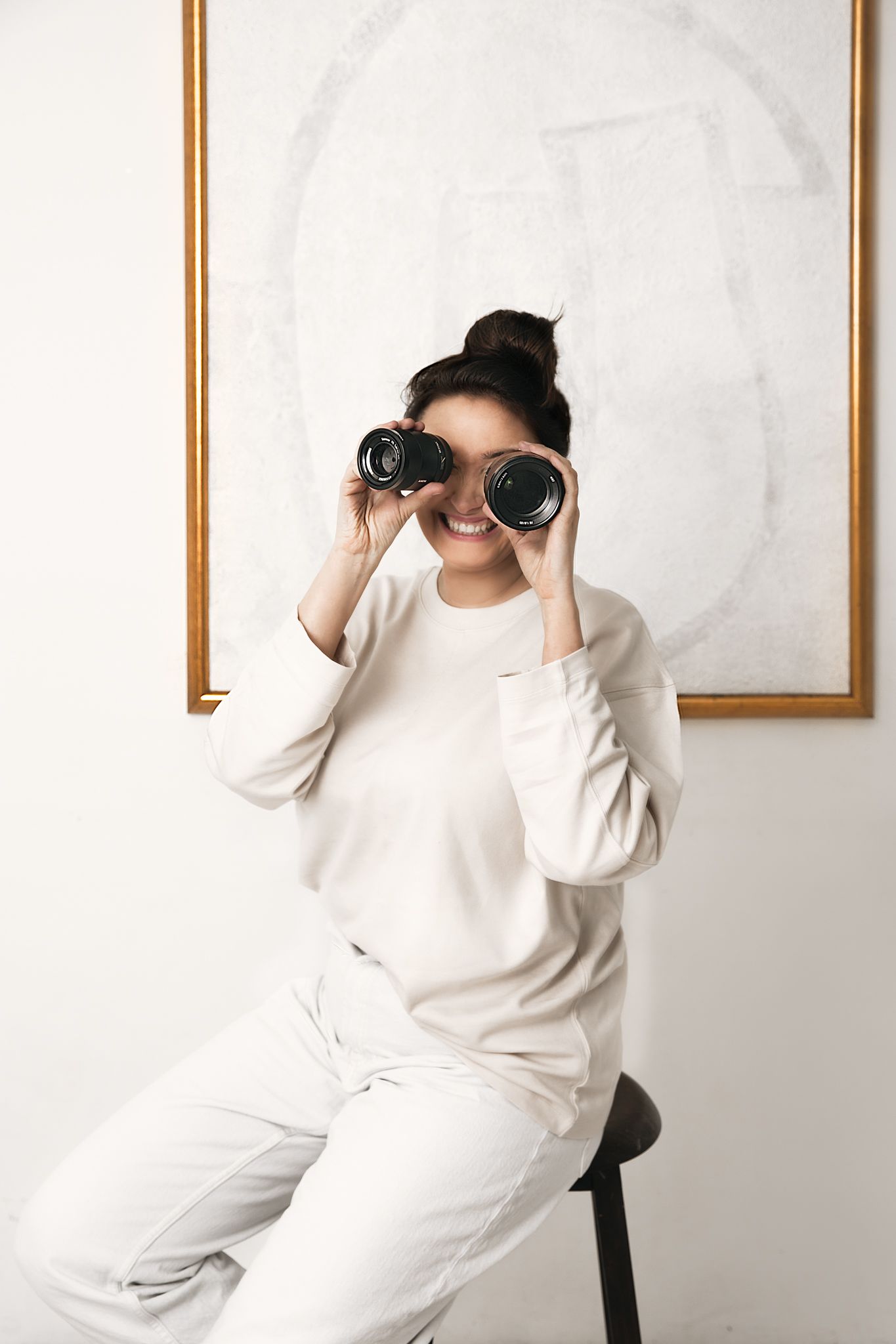 Person in white outfit looking through binoculars against white wall with framed art.