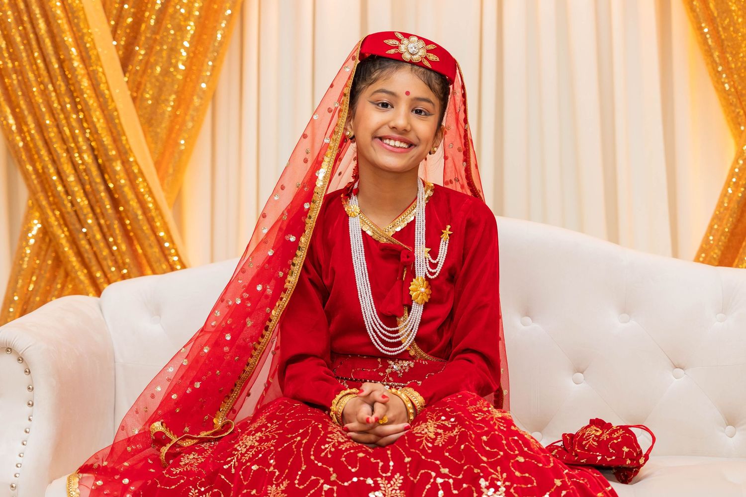 Traditional bride wearing red and gold attire sits on white couch against golden backdrop.