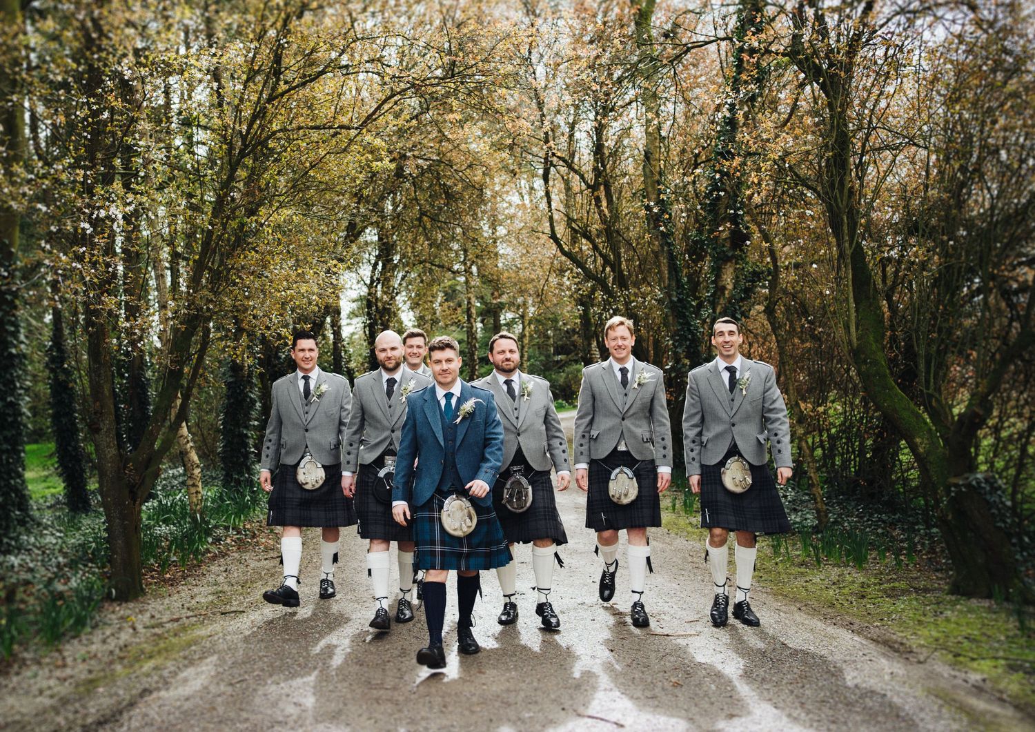 Groomsmen in traditional kilts walking together on a wet path surrounded by trees.