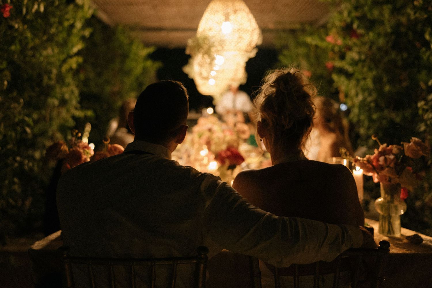 Romantic silhouette of a couple sitting together at a candlelit outdoor wedding reception dinner table.